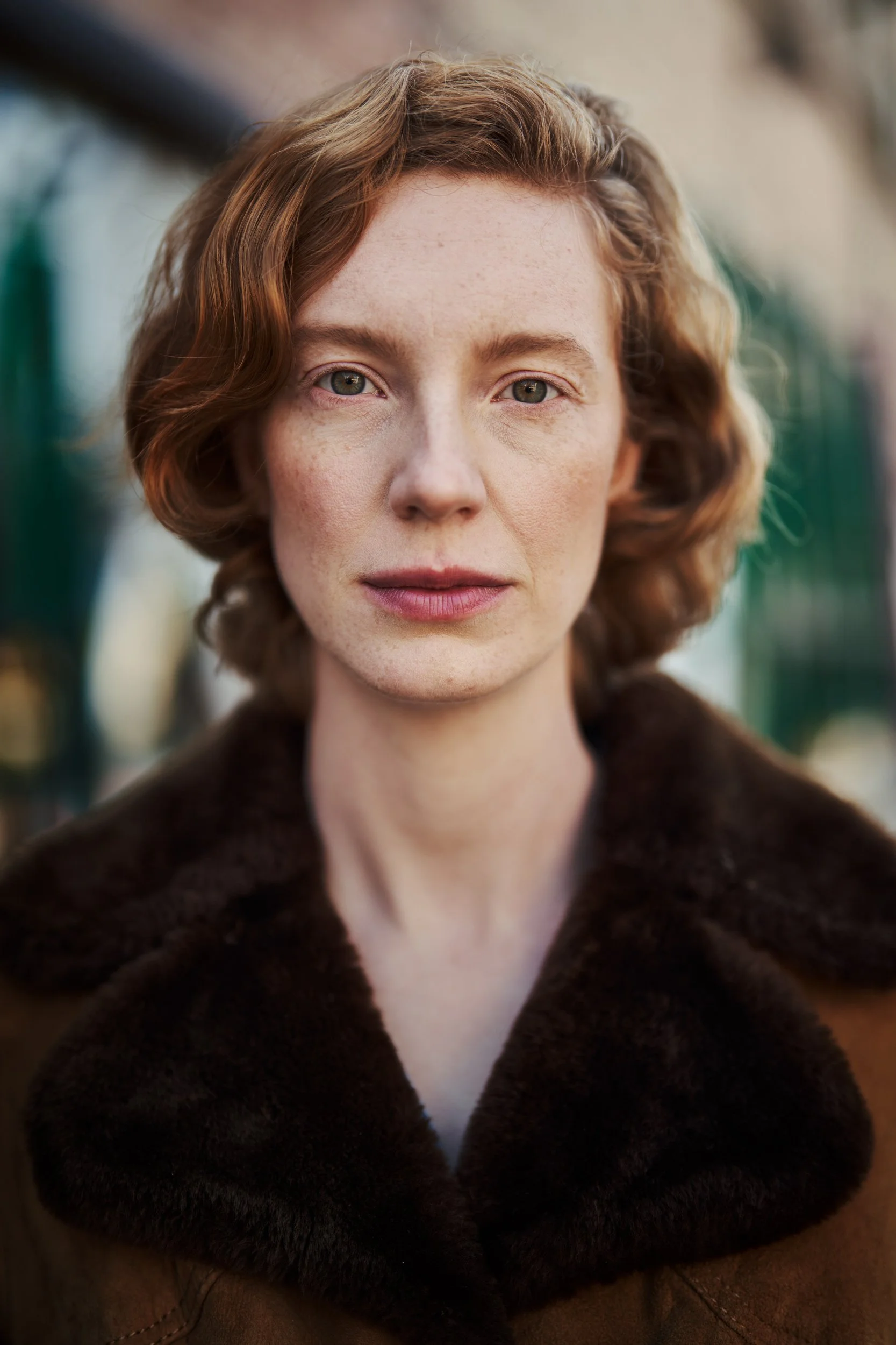 Headshot der Schauspielerin Luise Wolfram im braunen Mantel mit dunklem Fellkragen und direktem Blick vor unscharfem Straßenhintergrund, fotografiert von Koszki Photography Berlin.