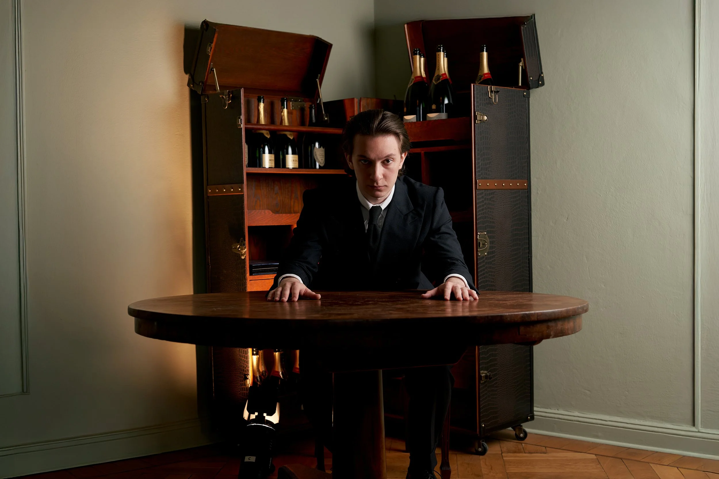 Porträt von Roland Junghals, Komponist und Musiker, aufgenommen in der Location Maximilian Mogg. Er sitzt formell gekleidet an einem runden Holztisch, beide Hände auf der Tischplatte. Hinter ihm steht ein geöffneter, elegant wirkender Schrankkoffer m