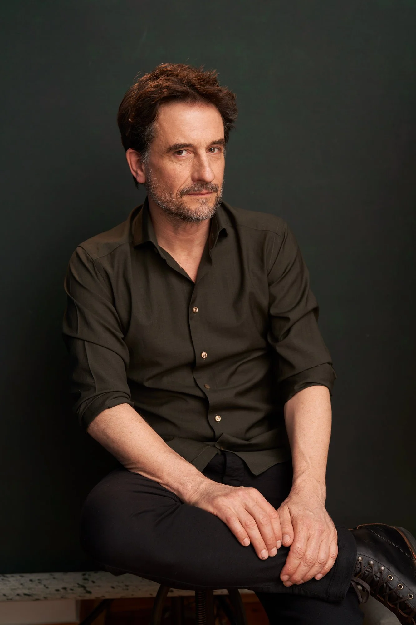 Studioporträt des Schauspielers Oliver Mommsen in olivgrünem Hemd, auf einem Hocker sitzend, die Hände auf dem Knie, vor dunkelgrünem Hintergrund, leicht geneigter Blick in die Kamera, fotografiert von Koszki Photography Berlin.