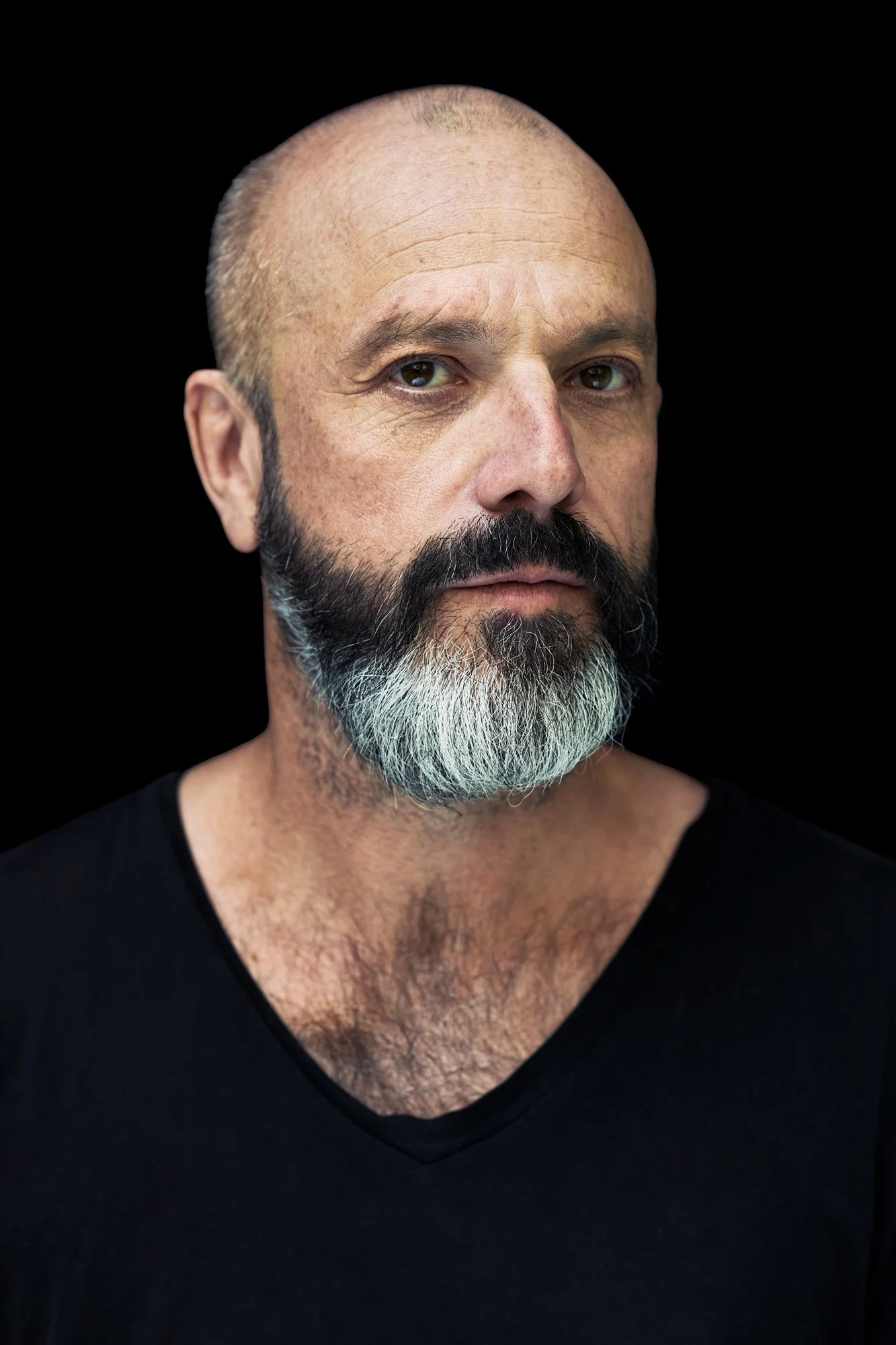 Porträt von Jan Plewka, Sänger der Rockband Selig, mit rasiertem Kopf, schwarz-grauem Vollbart und schwarzem V-Shirt, mit ernstem Blick vor schwarzem Hintergrund, fotografiert von Koszki Photography Berlin.