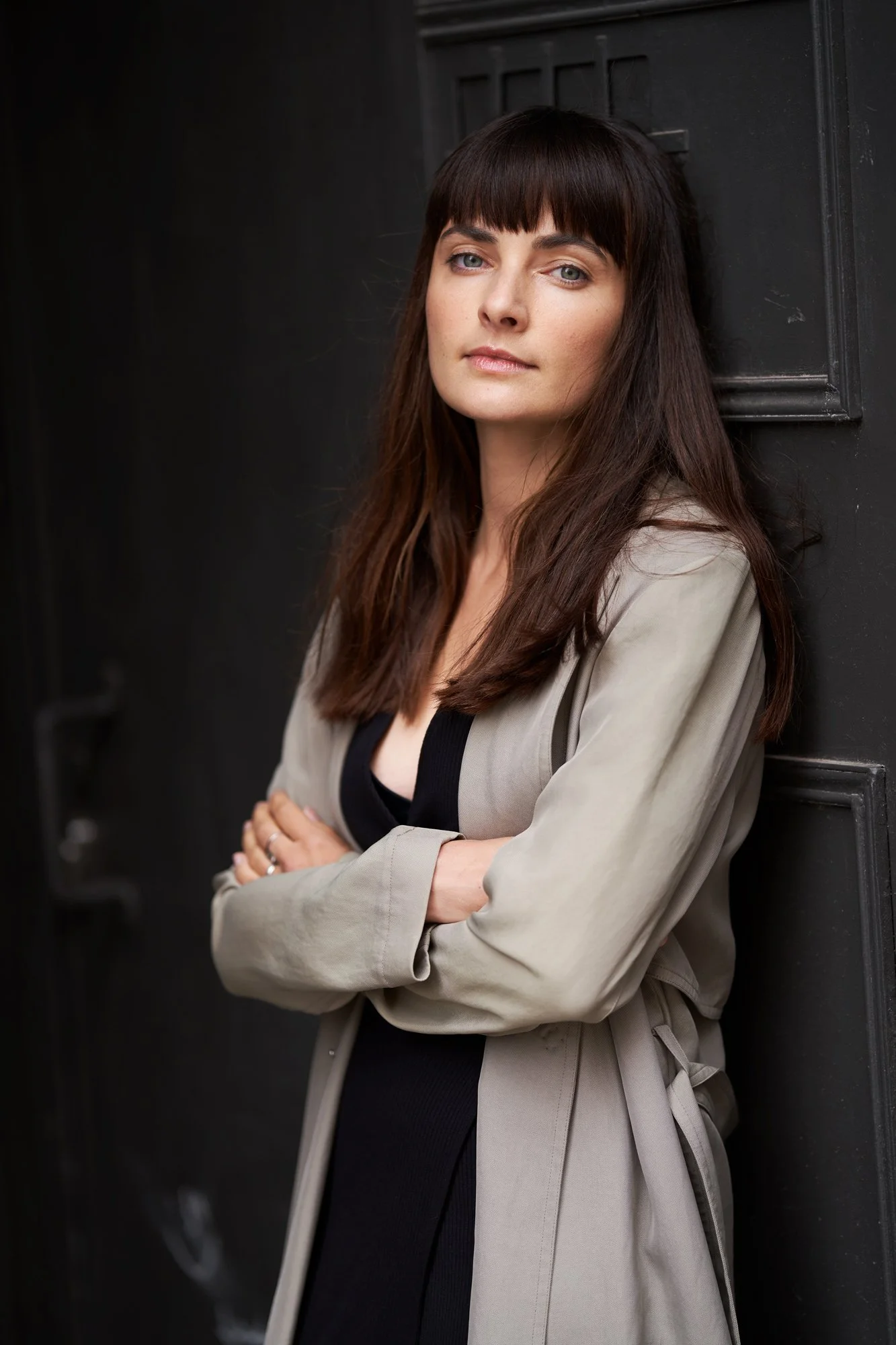 Porträtaufnahme der Schauspielerin Paula Schramm mit langem dunklem Haar und Pony im beigen Trenchcoat über schwarzem Kleid, mit verschränkten Armen an eine schwarze Holztür gelehnt, ernster Blick in die Kamera, fotografiert von Koszki Photography Be