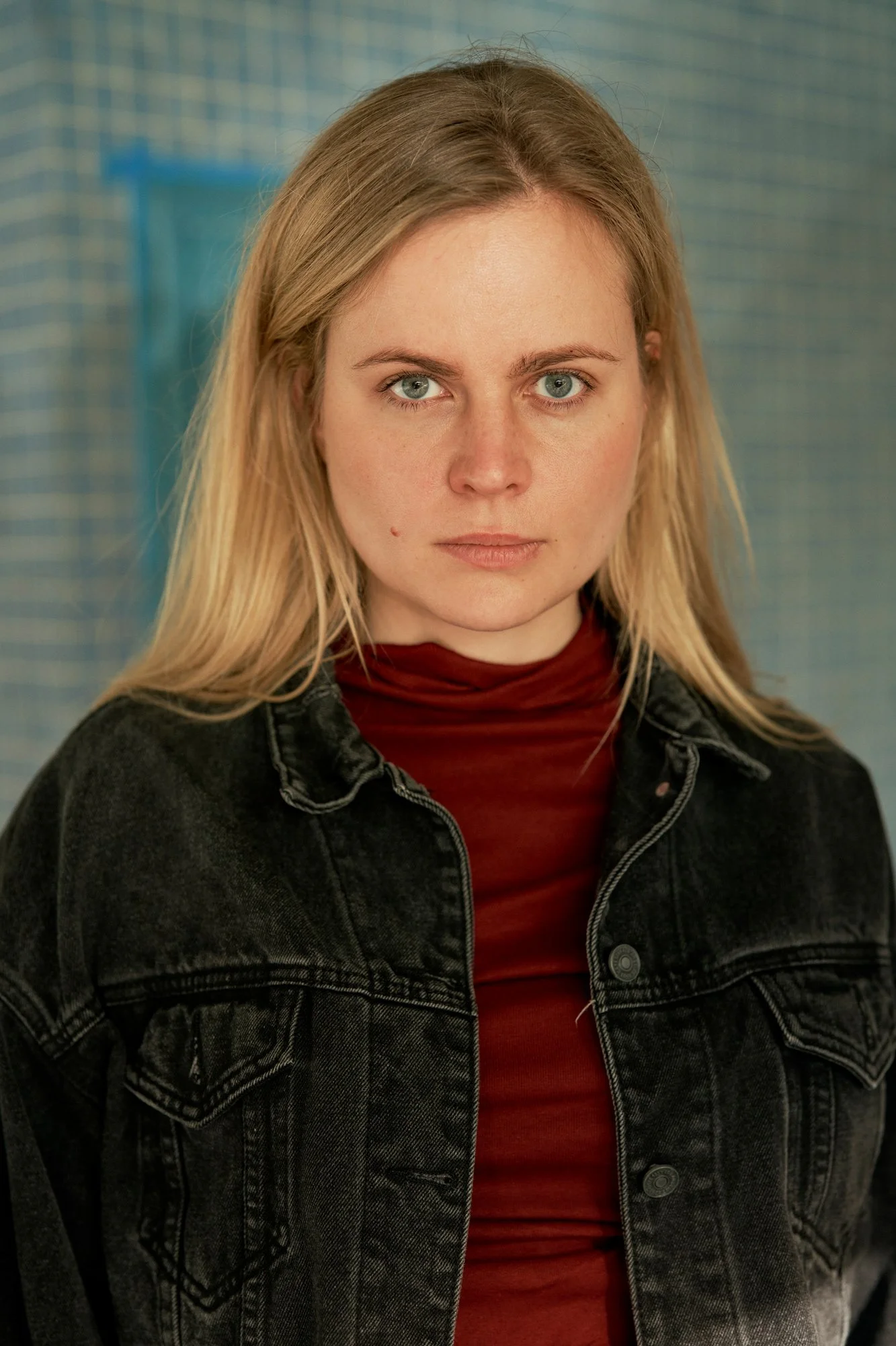 Headshot der Schauspielerin und Sängerin Kara Edenberg mit langen blonden Haaren in schwarzer Jeansjacke über rotem Rollkragenpullover, frontal mit ernstem Blick in die Kamera vor einer blauen Kachelwand, fotografiert von Koszki Photography Berlin.