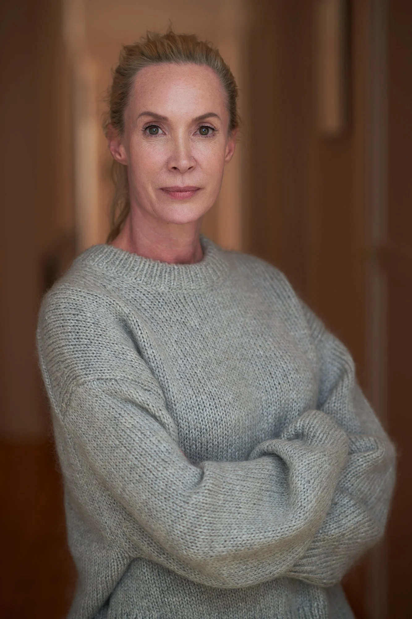 Porträt der Regisseurin und Schauspielerin Feo Aladag im grauen Strickpullover mit verschränkten Armen und zurückgebundenen Haaren, mit ruhigem Blick in die Kamera vor warmem braunem Hintergrund, fotografiert von Koszki Photography Berlin.