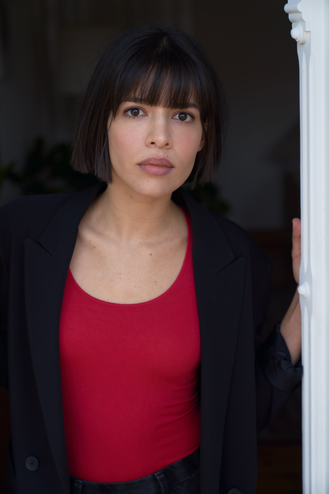 Porträt der Schauspielerin Jasmin Lord im schwarzen Blazer über rotem Top, die Hand an einem weißen Türrahmen, mit direktem Blick in die Kamera vor dunklem Hintergrund, fotografiert von Koszki Photography Berlin.