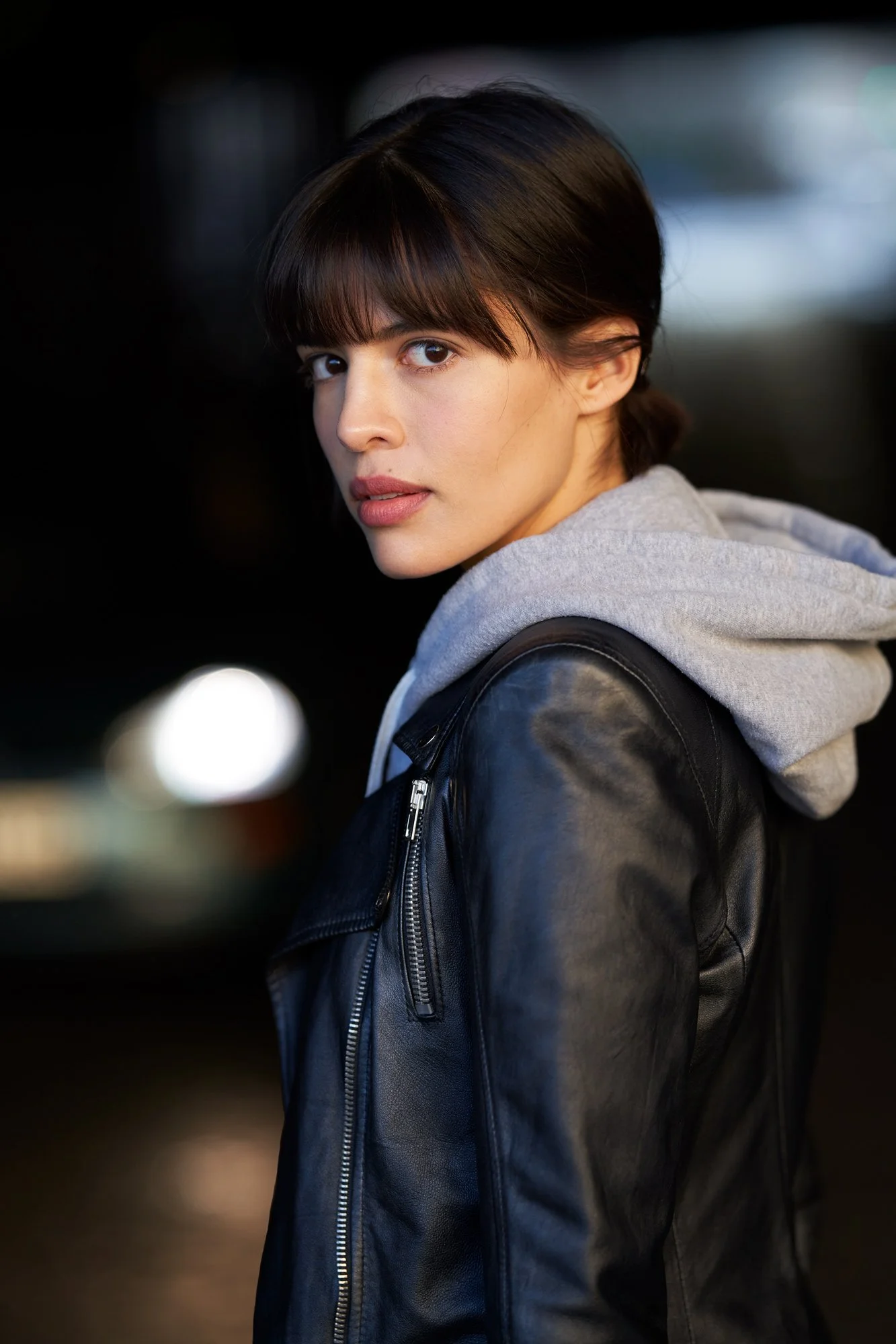 Porträt der Schauspielerin Jasmin Lord in schwarzer Lederjacke über grauem Kapuzenpullover, mit Blick über die Schulter in die Kamera vor dunklem Hintergrund mit Lichtreflexen, fotografiert von Koszki Photography Berlin.