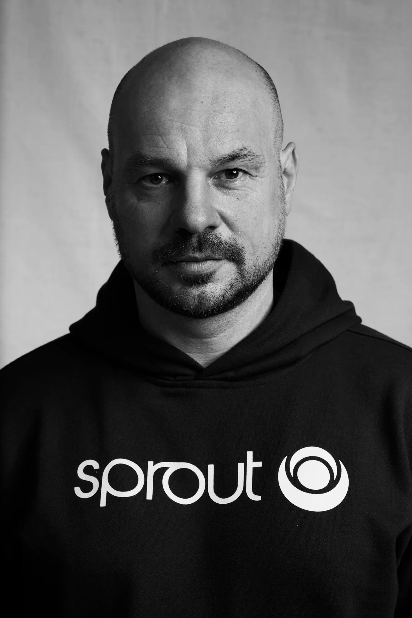 Schwarz-Weiß-Porträt des DJs und Produzenten D-Nox im schwarzen Sprout-Hoodie mit weißem Logo und ernstem, direktem Blick vor hellem Hintergrund, fotografiert von Koszki Photography Berlin.