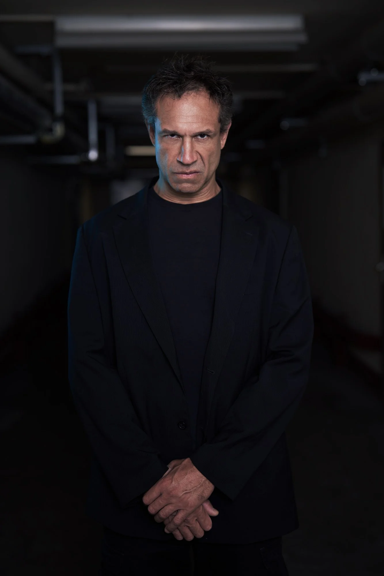 Porträtaufnahme von Evil Jared Hasselhoff (Bloodhound Gang) in schwarzem Blazer und Shirt, stehend in einem dunklen Kellergang mit gefalteten Händen, kühles Seitenlicht, düsterer Blick in die Kamera, fotografiert von Koszki Photography Berlin.