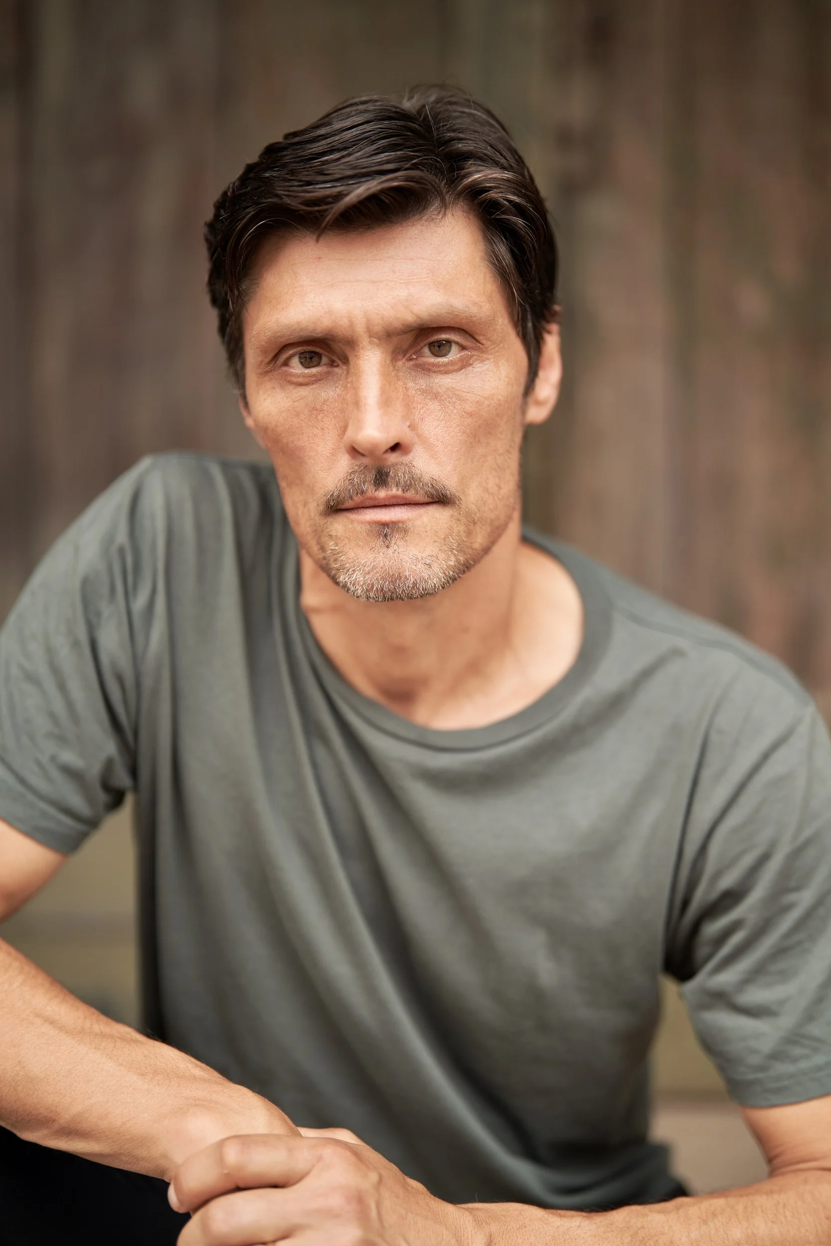 Headshot des Schauspielers Stipe Erceg im graugrünen T-Shirt mit aufgestützten Armen und intensivem Blick vor einer Holzwand, fotografiert von Koszki Photography Berlin.