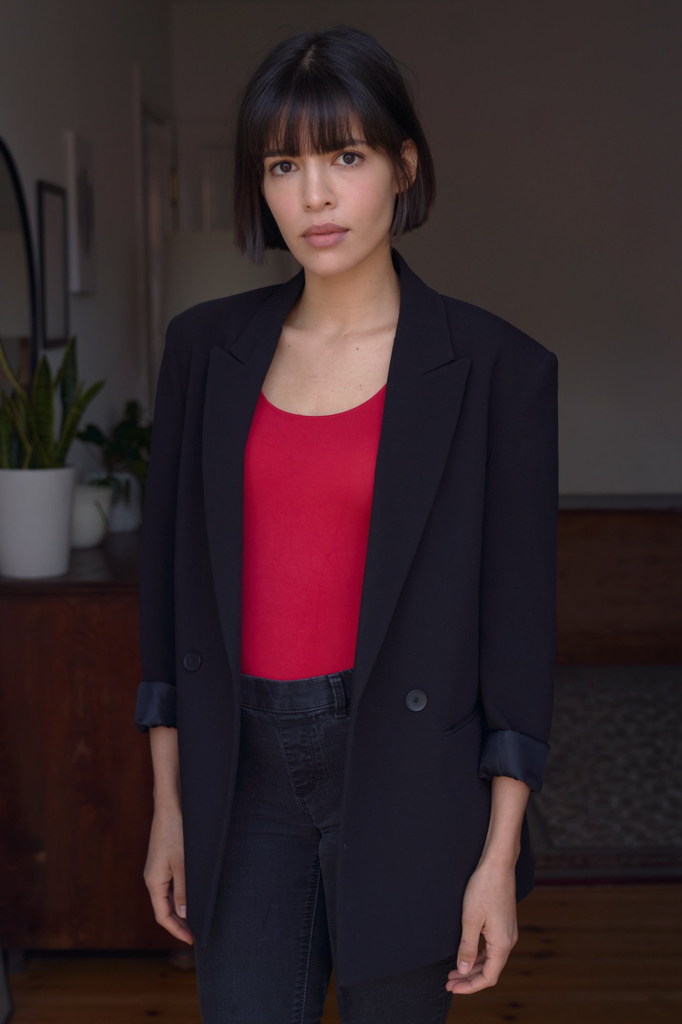 Porträt der Schauspielerin Jasmin Lord mit schwarzem Bob und Pony im schwarzen Blazer über rotem Top, mit ruhigem Blick in die Kamera in einem Wohnraum mit Zimmerpflanzen, fotografiert von Koszki Photography Berlin.