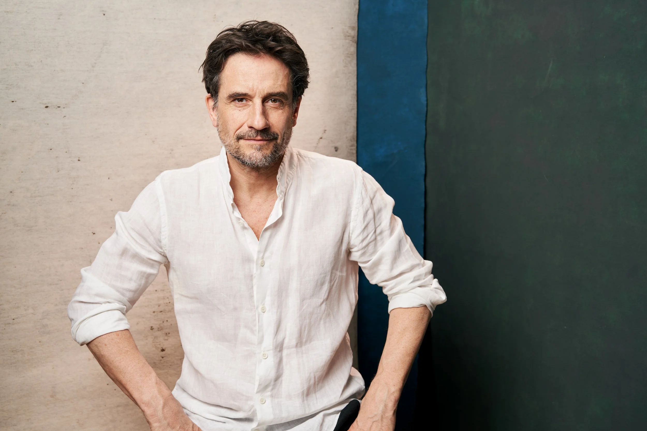 Studioporträt des Schauspielers Oliver Mommsen im weißen Leinenhemd mit hochgekrempelten Ärmeln, die Hände in die Hüften gestützt, vor beigem, blauem und dunkelgrünem Hintergrund, fotografiert von Koszki Photography Berlin.
