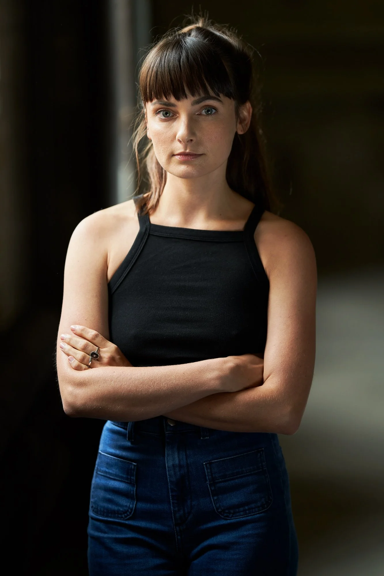 Porträtaufnahme der Schauspielerin Paula Schramm in schwarzem Top und Jeans mit verschränkten Armen, vor dunklem Hintergrund mit weichem Seitenlicht, direkter Blick in die Kamera, fotografiert von Koszki Photography Berlin.