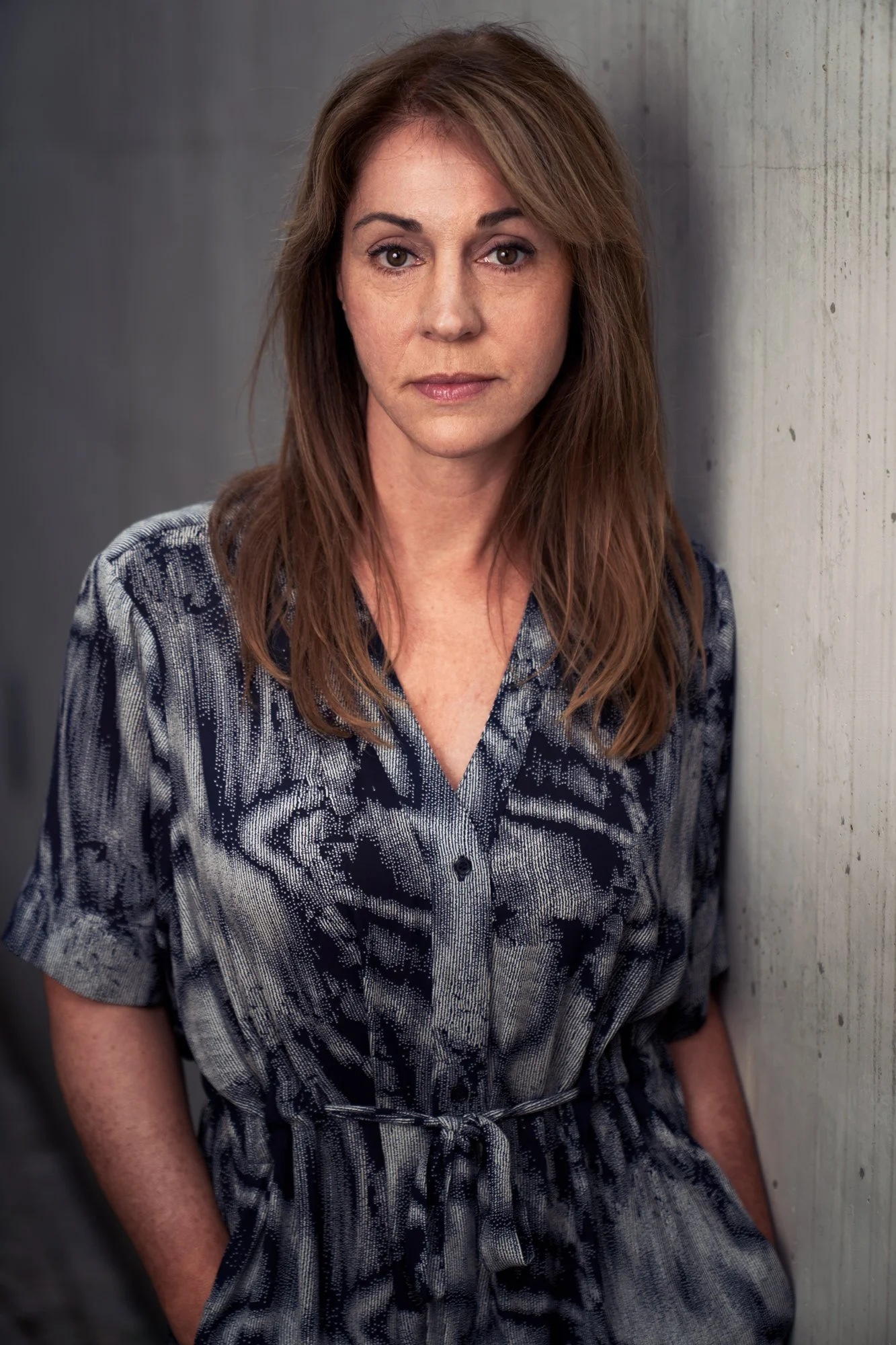 Porträtaufnahme der Schauspielerin Ulrike C. Tscharre in blau-grau gemustertem Kleid, an eine Betonwand gelehnt, ruhiger Blick in die Kamera, fotografiert von Koszki Photography Berlin.