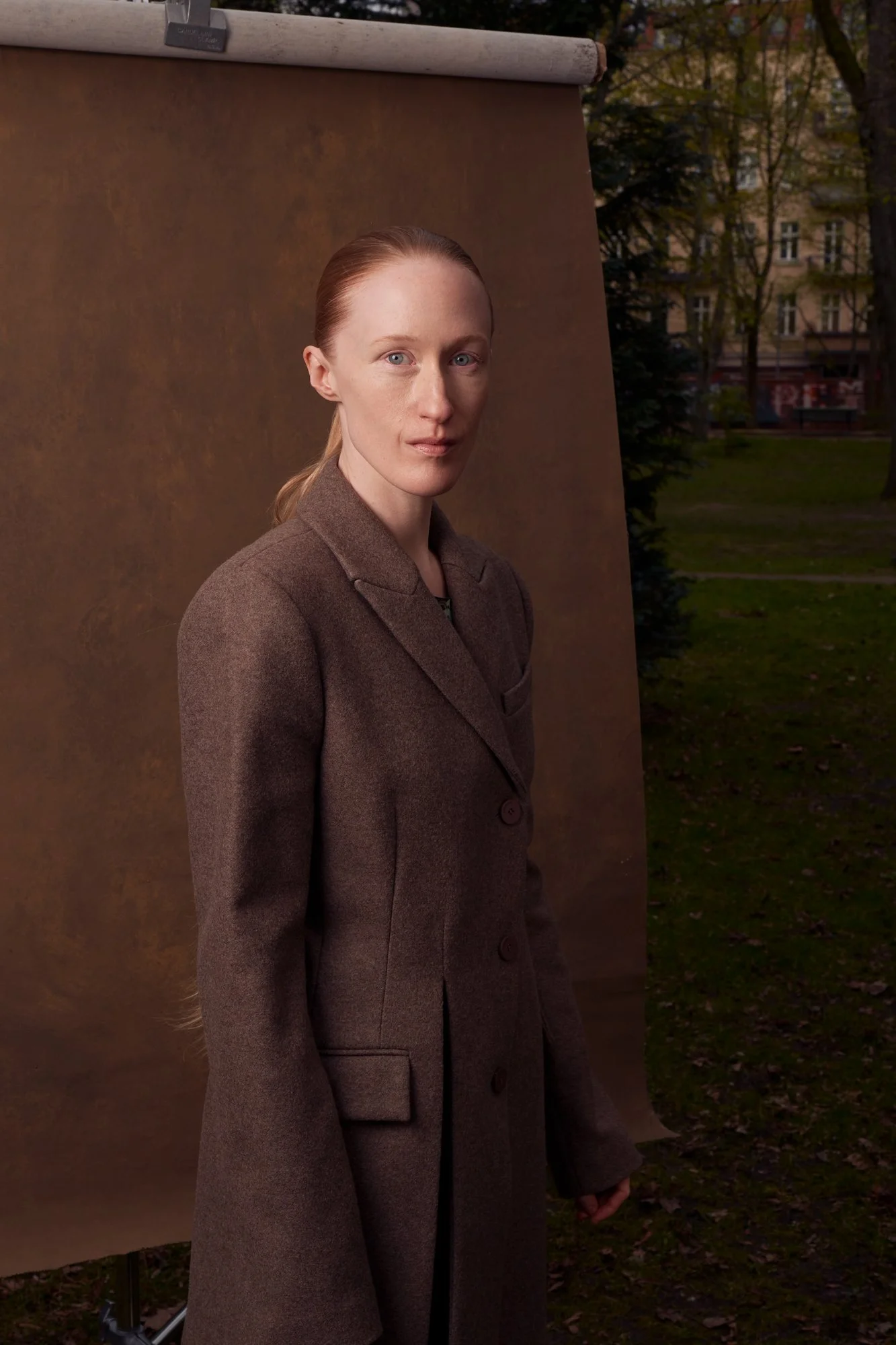 Porträt der Künstlerin und Schauspielerin Britta Thie mit nach hinten gebundenen Haaren im braunen Wollmantel, seitlich stehend mit direktem Blick in die Kamera vor einer braunen Hintergrundrolle in einem Berliner Park, fotografiert von Koszki Photog