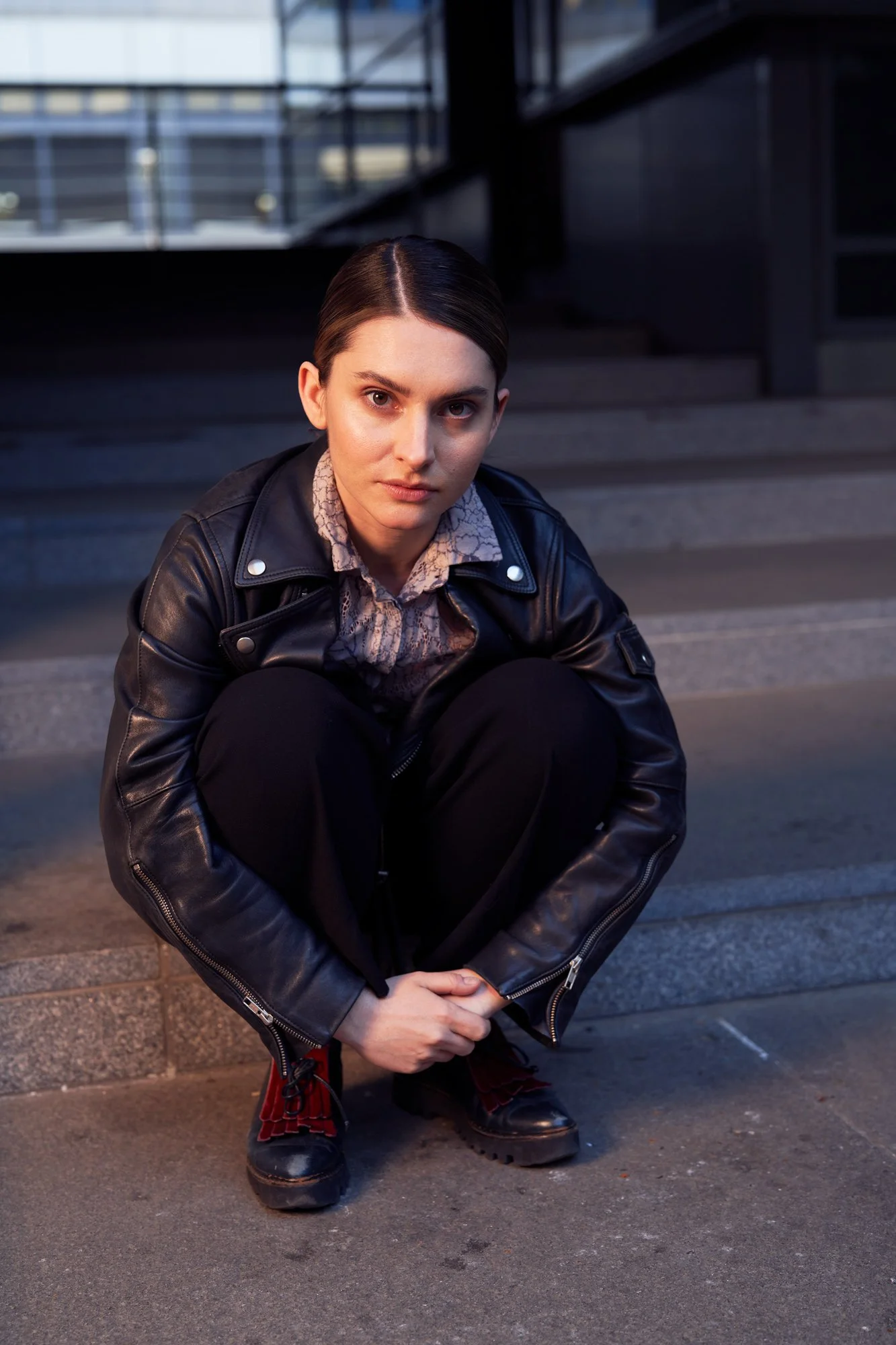 Porträtaufnahme der Schauspielerin Romina Küper mit zurückgekämmtem Haar in schwarzer Lederjacke und Spitzenbluse, in der Hocke auf einer Steintreppe vor moderner Architektur i