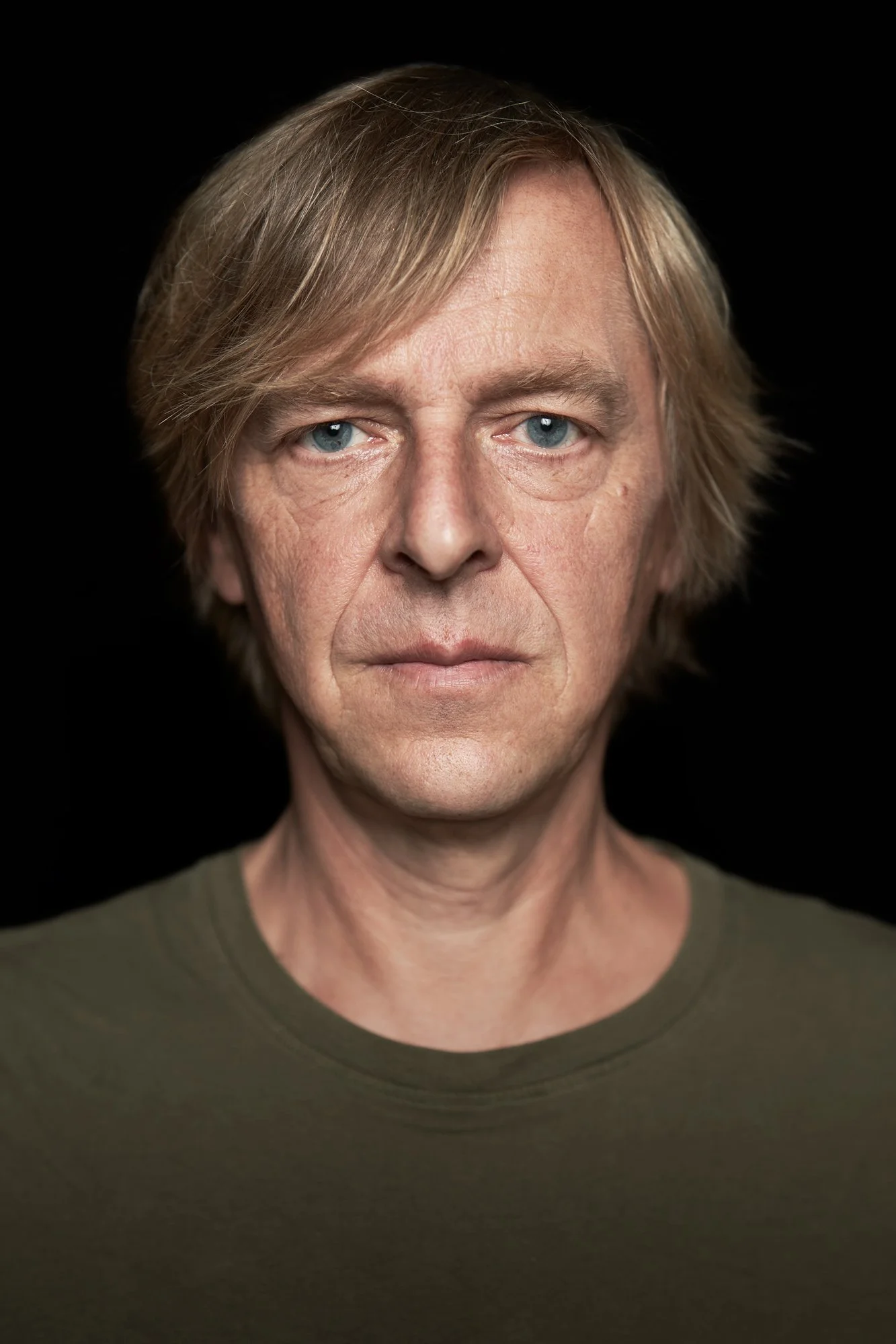 Porträt des Schlagzeugers Stephan „Stoppel“ Eggert von der Rockband Selig, im olivgrünen T-Shirt mit ernstem, direktem Blick vor schwarzem Hintergrund, fotografiert von Koszki Photography Berlin.