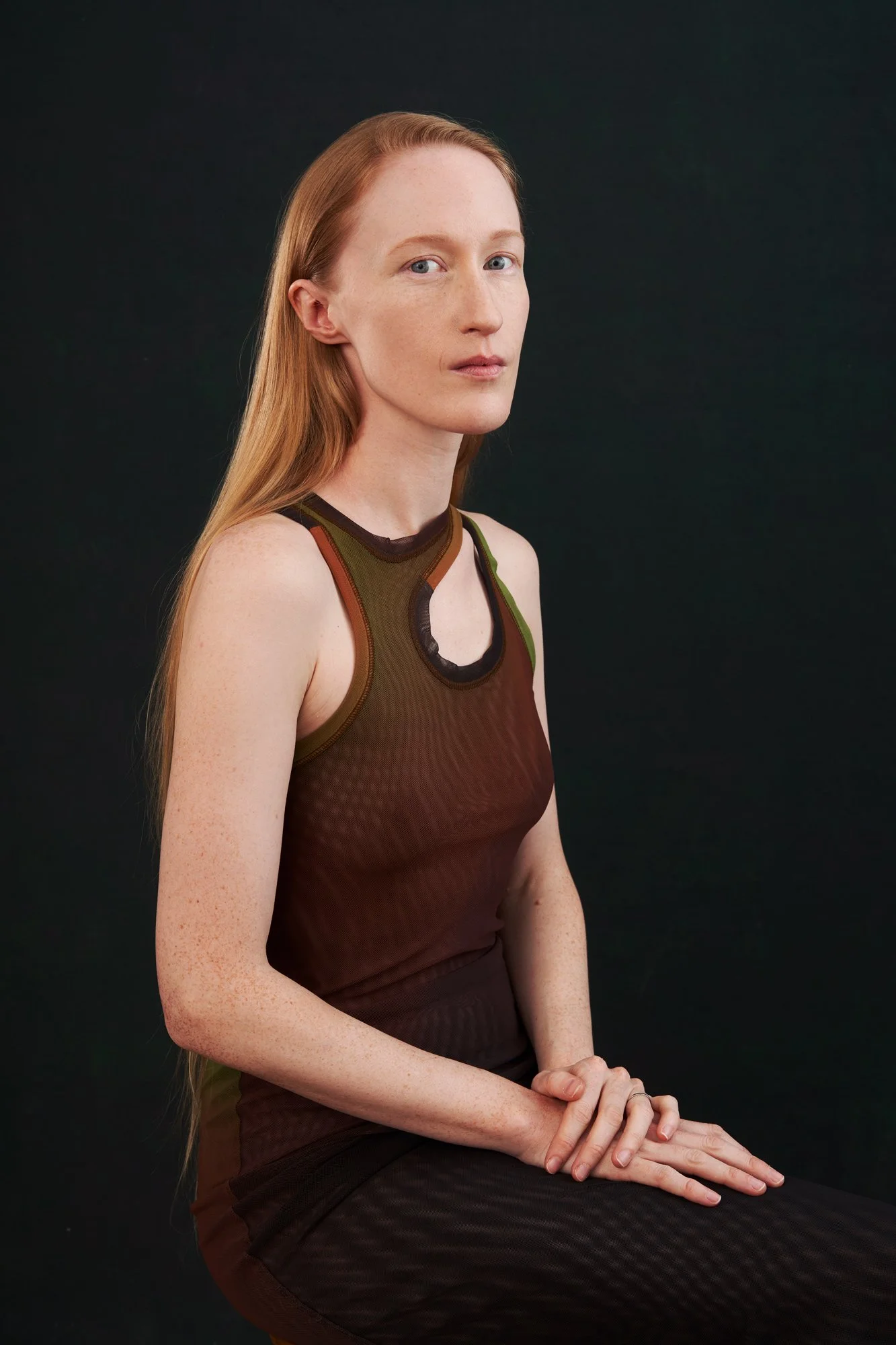 Studioportrait der Künstlerin und Schauspielerin Britta Thie mit langen roten Haaren im braun-grünen Mesh-Top, seitlich sitzend mit gefalteten Händen und Blick in die Kamera, vor dunklem Hintergrund mit gezielt gesetztem Licht, fotografiert von Koszk