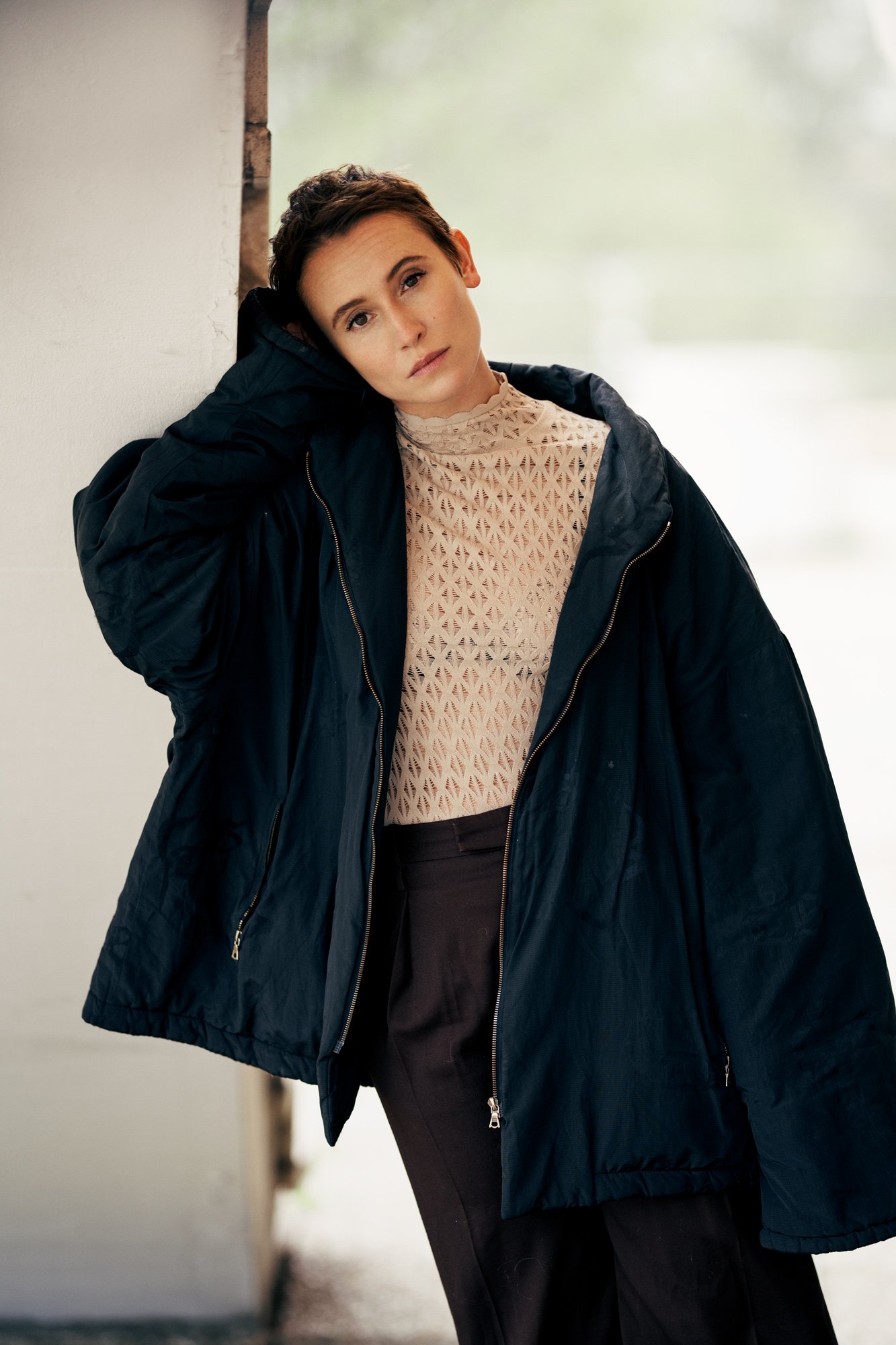 Porträtaufnahme der Schauspielerin Peri Baumeister in dunkelblauer Oversize-Jacke über beigem Lochmuster-Oberteil, an eine Säule gelehnt, Kopf geneigt, fotografiert von Koszki Photography Berlin.