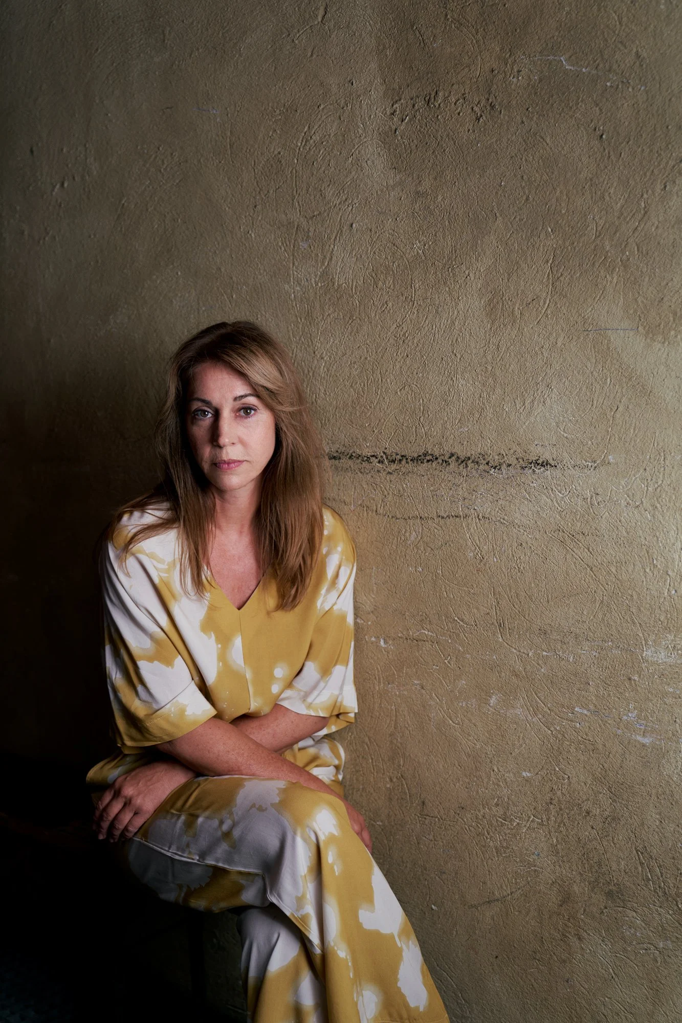 Porträtaufnahme der Schauspielerin Ulrike C. Tscharre im senfgelb-weiß gemusterten Kleid, sitzend vor einer strukturierten ockerfarbenen Wand, ruhiger Blick in die Kamera, fotografiert von Koszki Photography Berlin.