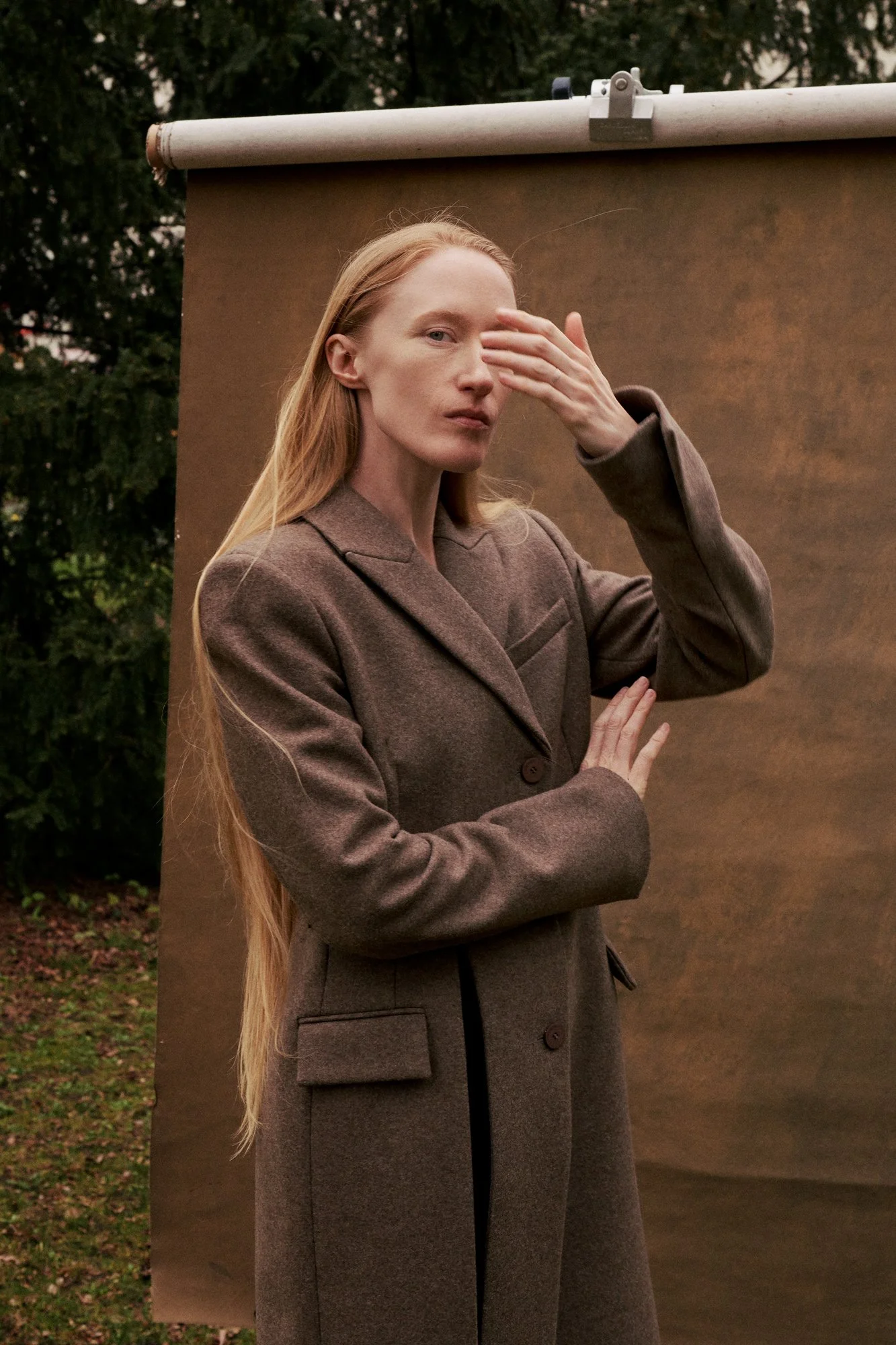 Porträt der Künstlerin und Schauspielerin Britta Thie mit langen blonden Haaren im braunen Wollmantel, die Hand vor dem Gesicht, vor einer aufgehängten braunen Fotohintergrundrolle im Freien mit Bäumen im Hintergrund, fotografiert von Koszki Photogra