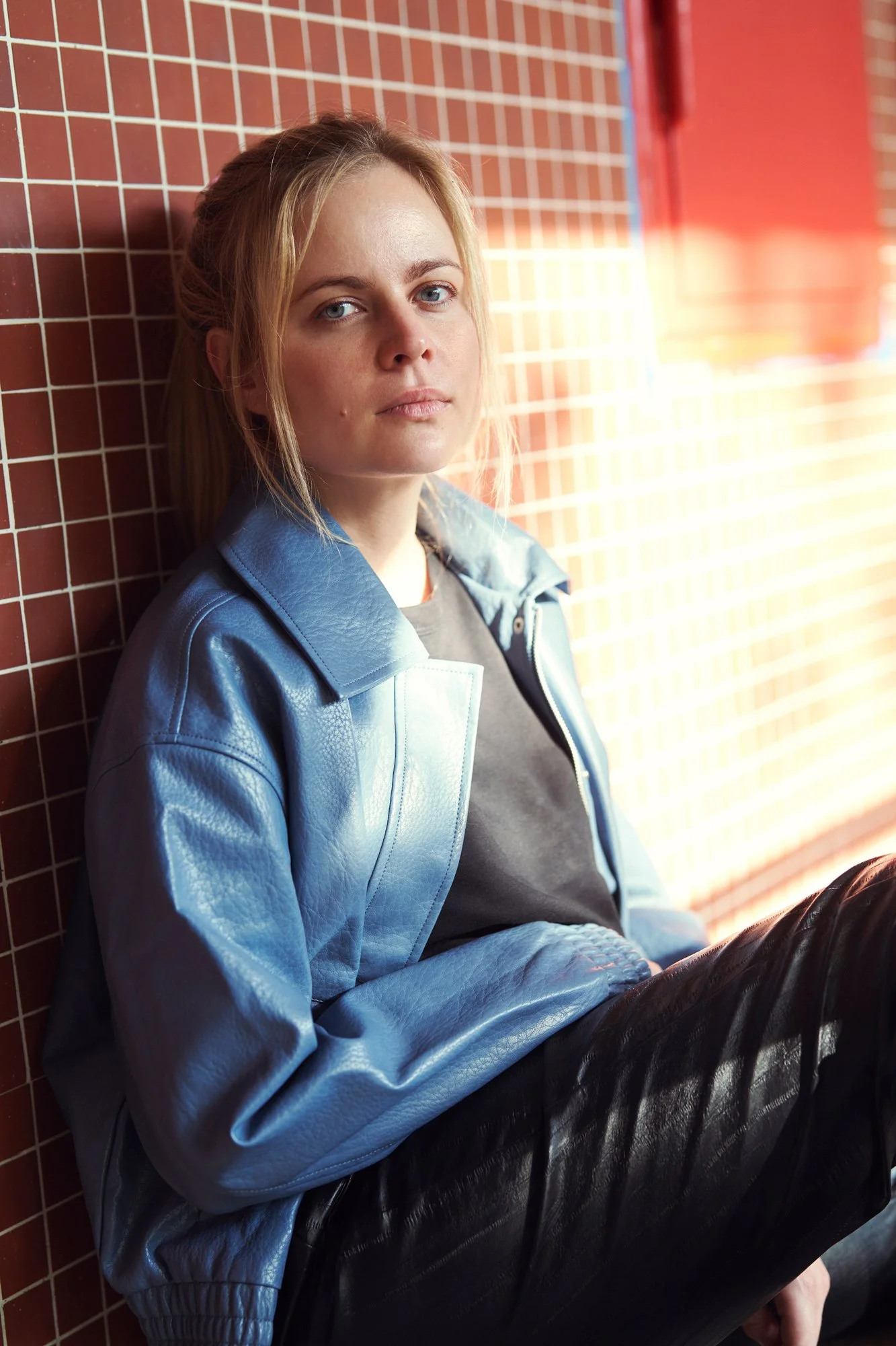 Porträt der Schauspielerin und Sängerin Kara Edenberg im blauen Lederblouson über grauem Pullover, an eine rote Kachelwand gelehnt, mit ruhigem Blick in die Kamera im warmen Gegenlicht, fotografiert von Koszki Photography Berlin.