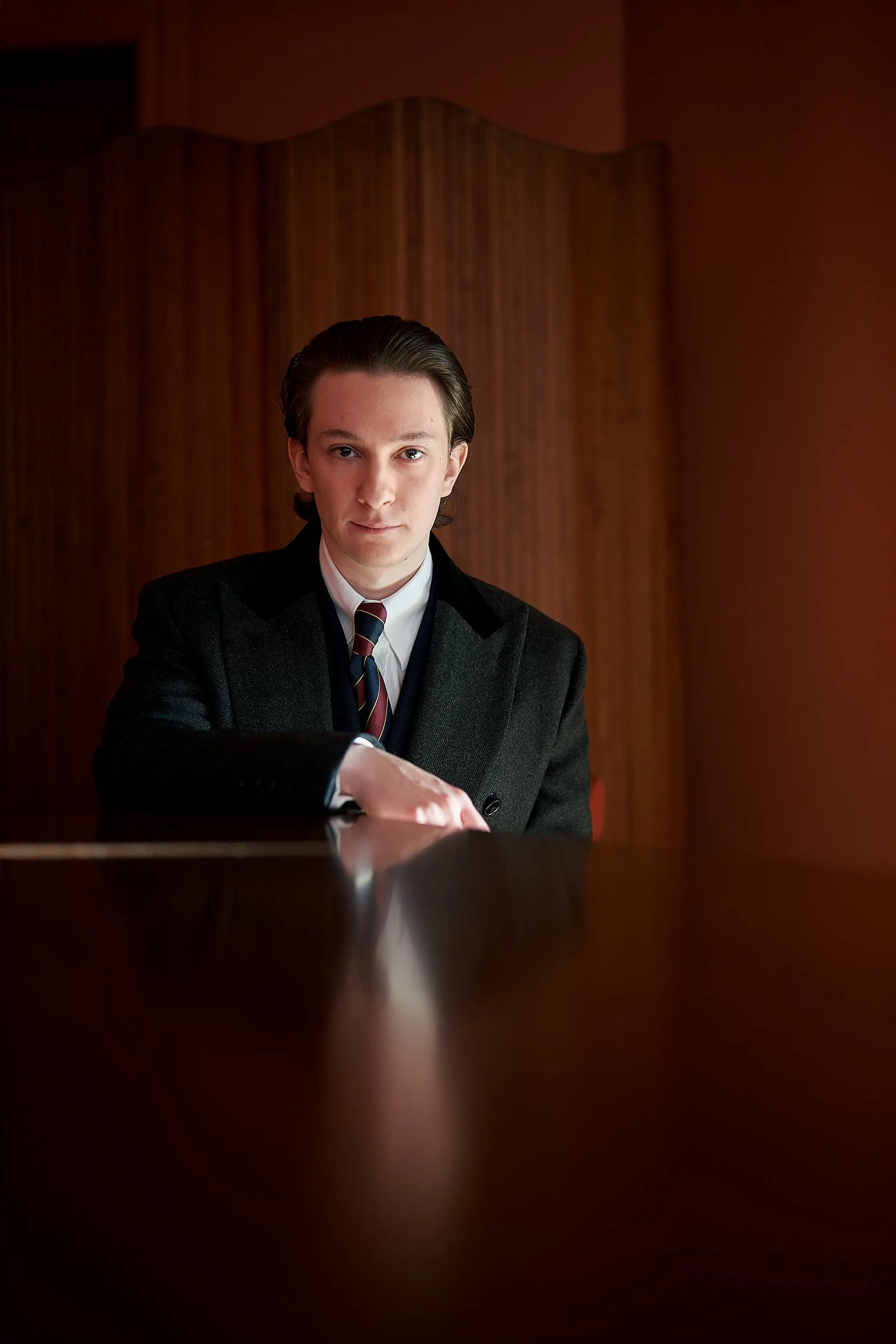 Stilvolles Porträt eines Mannes im dunklen Anzug an einem Holztisch, in warmem Licht. Die Szene erinnert an die elegante Ästhetik von Maximilian Mogg und trägt eine jazzige, komponistenhafte Stimmung. Keywords: Roland Junghals, Komponist, Jazz, Mann 