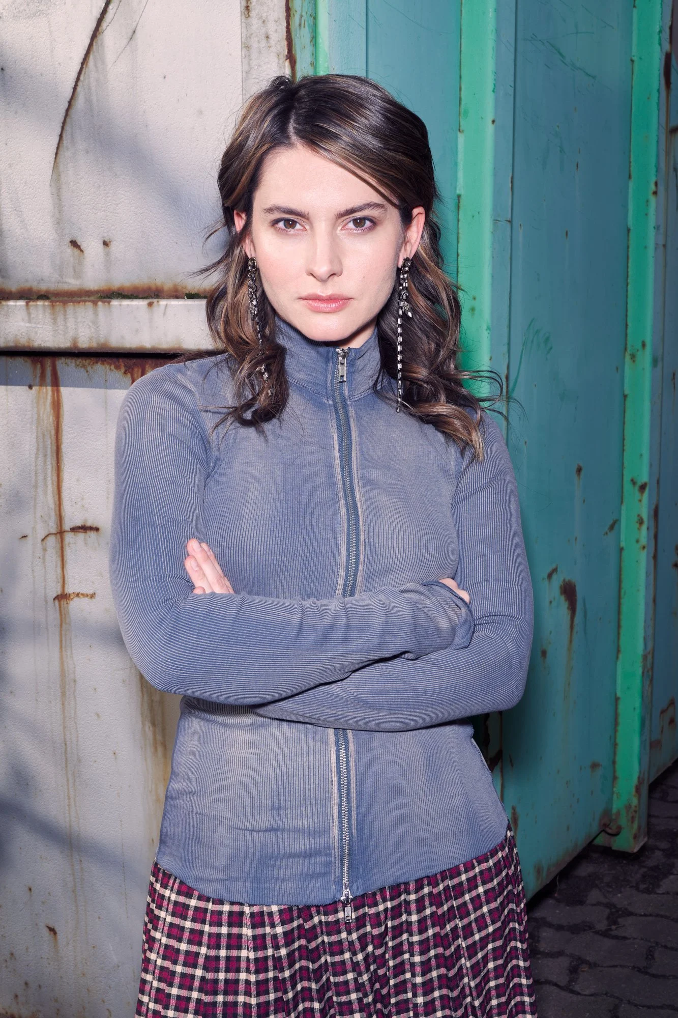 Porträtaufnahme der Schauspielerin Romina Küper mit welligem dunklem Haar, langen Ohrringen und grauer Zipp-Strickjacke, mit verschränkten Armen vor einem türkisen Container, im harten Licht, ernster Blick in die Kamera, fotografiert von Koszki Photo