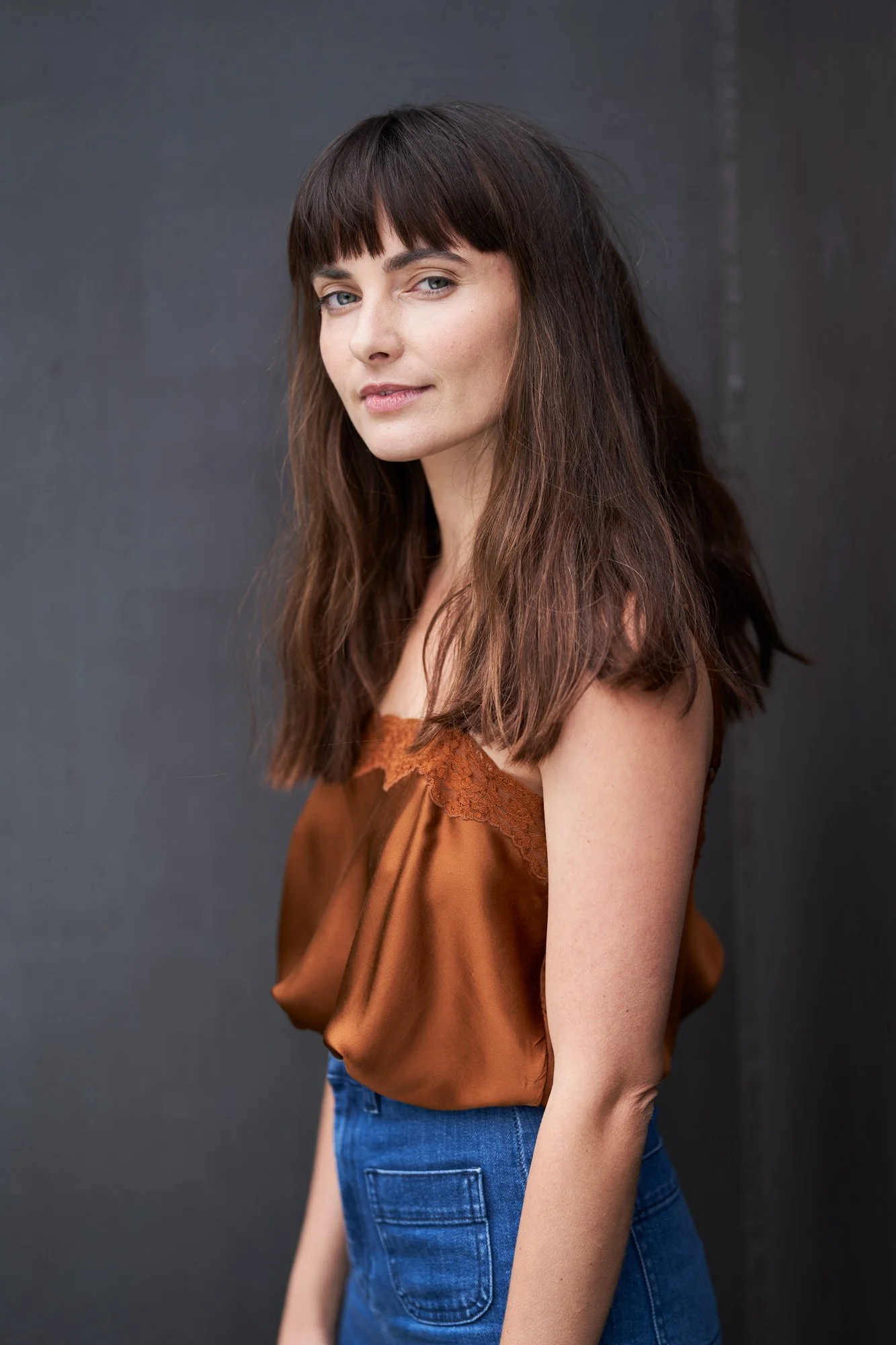 Porträtaufnahme der Schauspielerin Paula Schramm mit langem dunklem Haar und Pony im kupferfarbenen Seidentop mit Spitze und Jeans, seitlich stehend, Blick über die Schulter in die Kamera, fotografiert von Koszki Photography Berlin.