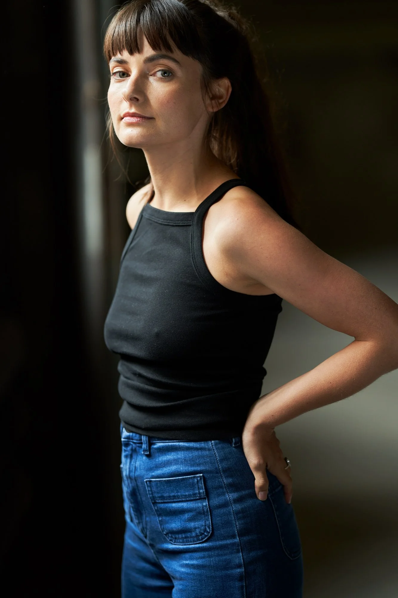 Porträtaufnahme der Schauspielerin Paula Schramm im schwarzen Top und Jeans, im Profil stehend mit der Hand an der Hüfte, Blick über die Schulter in die Kamera, vor dunklem Hintergrund mit weichem Seitenlicht, fotografiert von Koszki Photography Berl