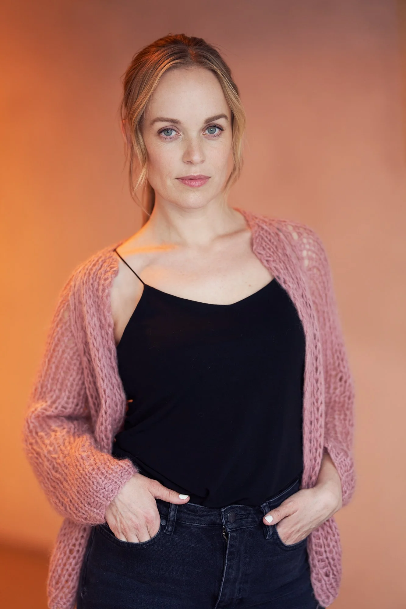Porträtaufnahme der Schauspielerin Friederike Kempter in rosa Strickjacke und schwarzem Top vor orangefarbenem Hintergrund, fotografiert von Koszki Photography Berlin.