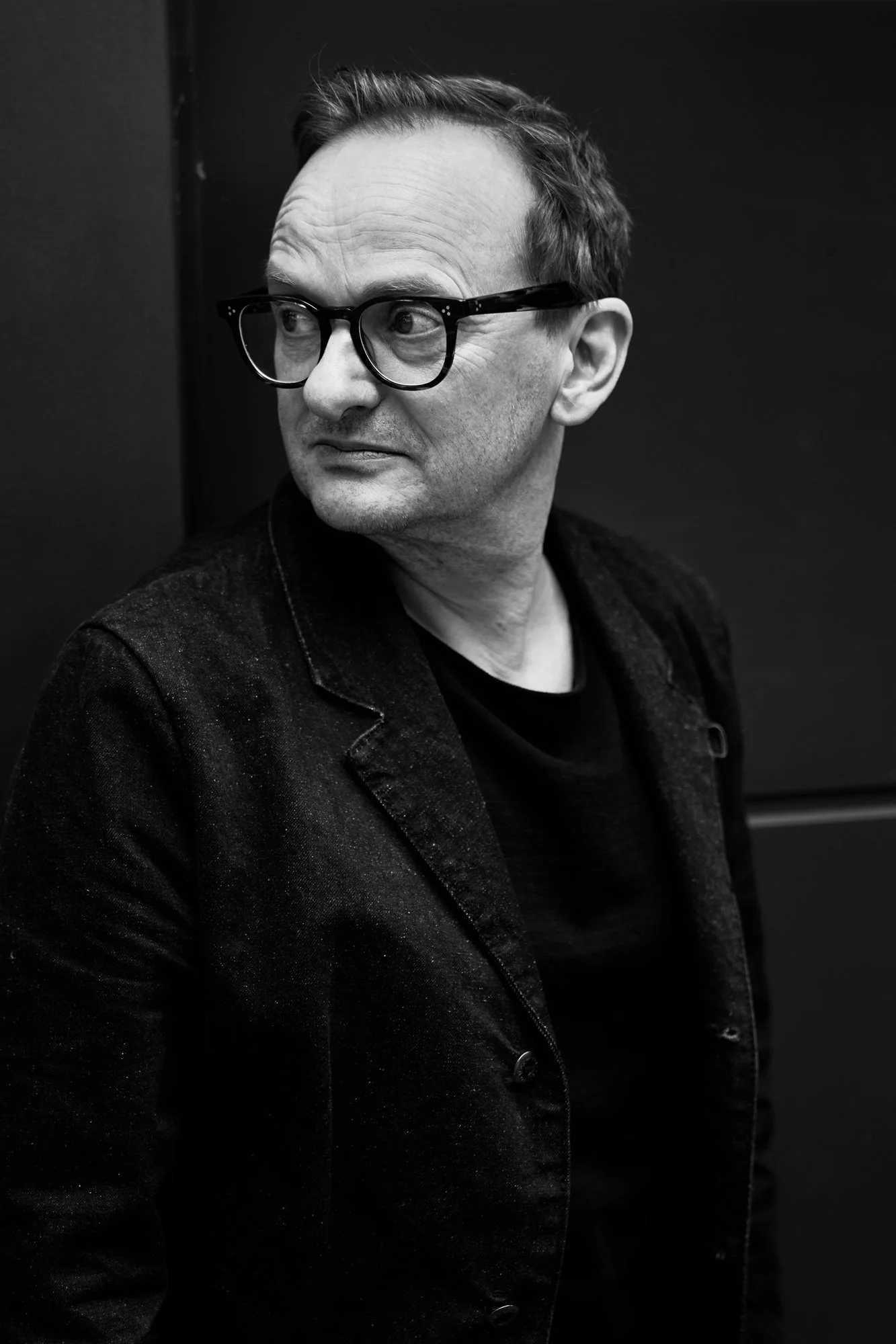 Schwarz-Weiß-Porträt des Schauspielers Milan Peschel mit Brille im dunklen Jeansblazer vor dunklem Hintergrund, Blick zur Seite, markanter Gesichtsausdruck, fotografiert von Koszki Photography Berlin.