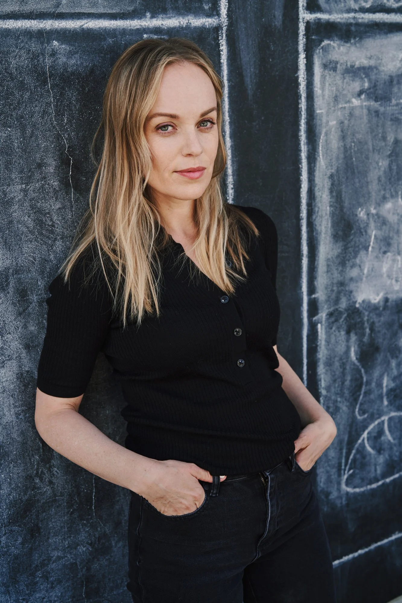 Porträtaufnahme der Schauspielerin Friederike Kempter in schwarzem Shirt und Jeans, an eine Tafelwand gelehnt, fotografiert von Koszki Photography Berlin.