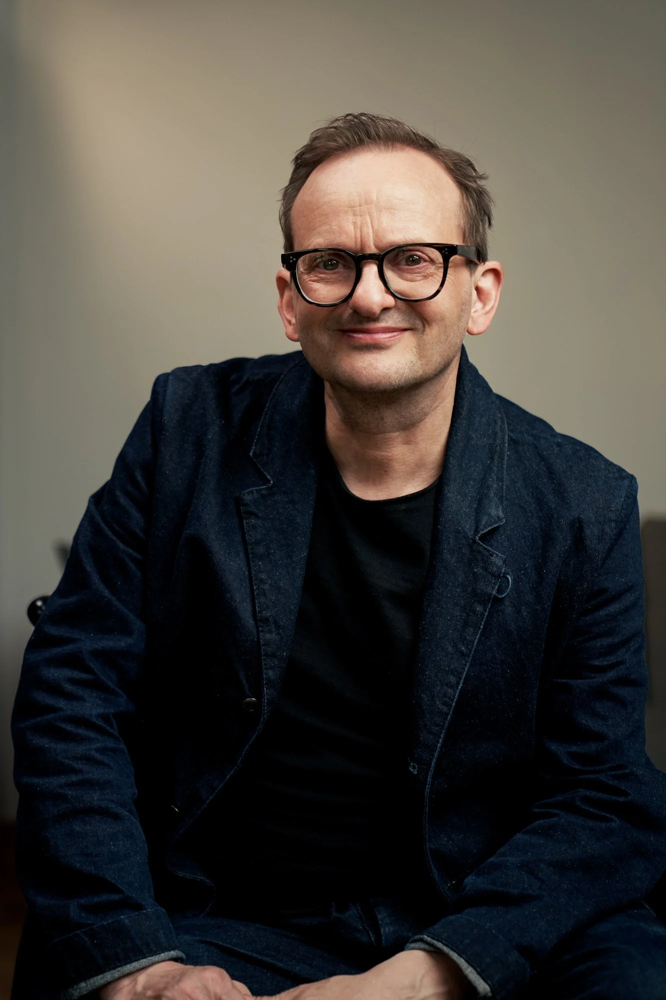 Porträtaufnahme des Schauspielers Milan Peschel mit Brille im dunkelblauen Jeansblazer und schwarzem Shirt, sitzend vor warmem beigem Hintergrund, freundliches Lächeln, fotografiert von Koszki Photography Berlin.
