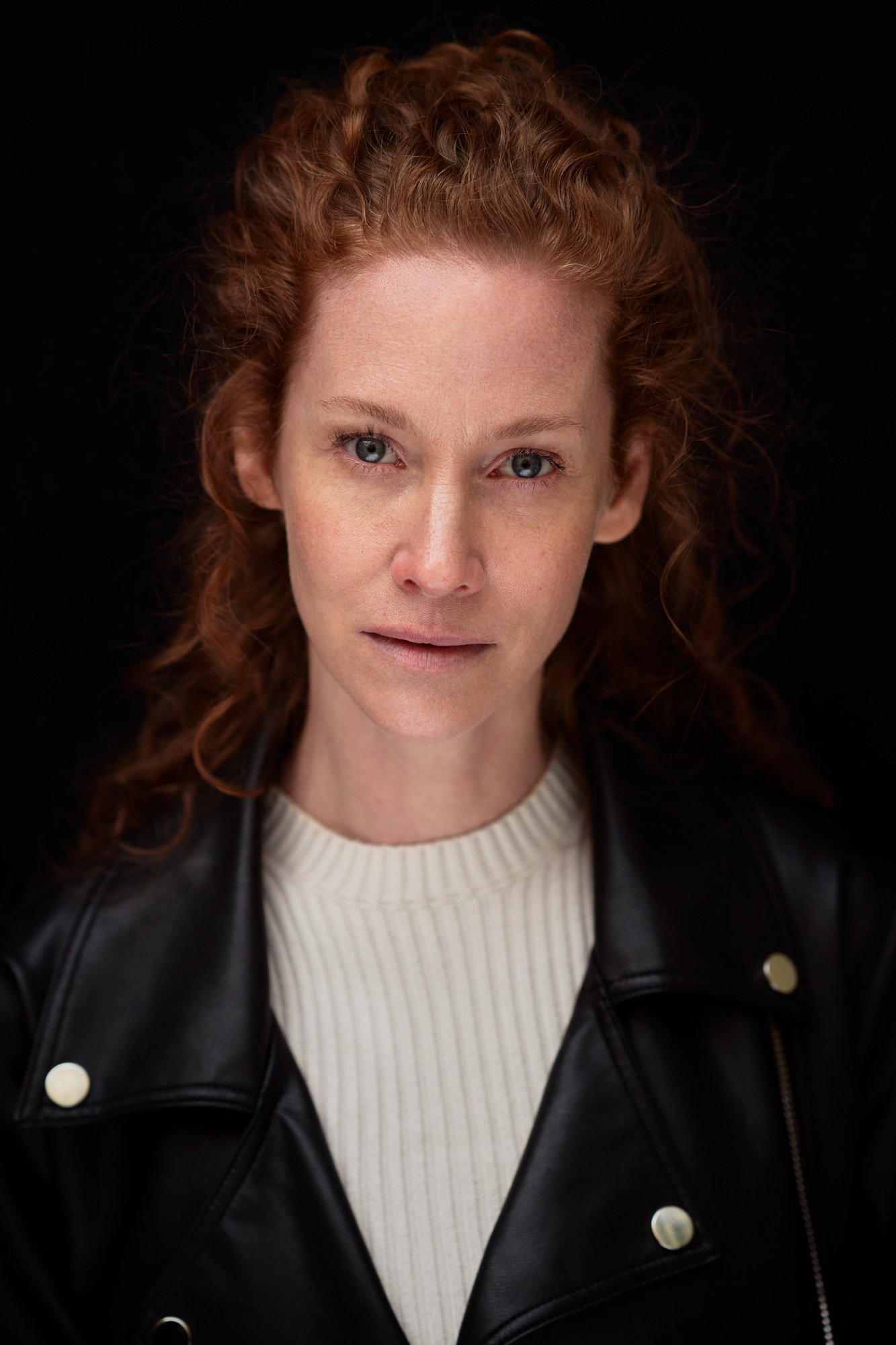 Headshot der Schauspielerin Sina-Valeska Jung mit lockigem rotem Haar in schwarzer Lederjacke und weißem Rippstrick-Pullover vor schwarzem Hintergrund, direkter Blick in die Kamera, fotografiert von Koszki Photography Berlin.