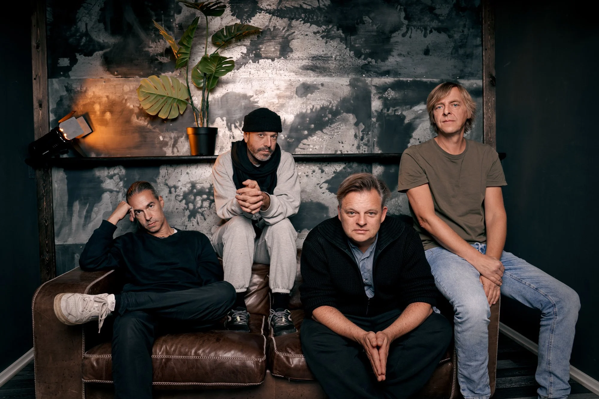 Bandfoto der Rockband Selig mit Sänger Jan Plewka, vier Musiker auf einem braunen Ledersofa vor einer metallisch gemusterten Wand mit Monstera-Pflanze im Backstage des Huxleys Neue Welt Berlin, fotografiert von Koszki Photography.