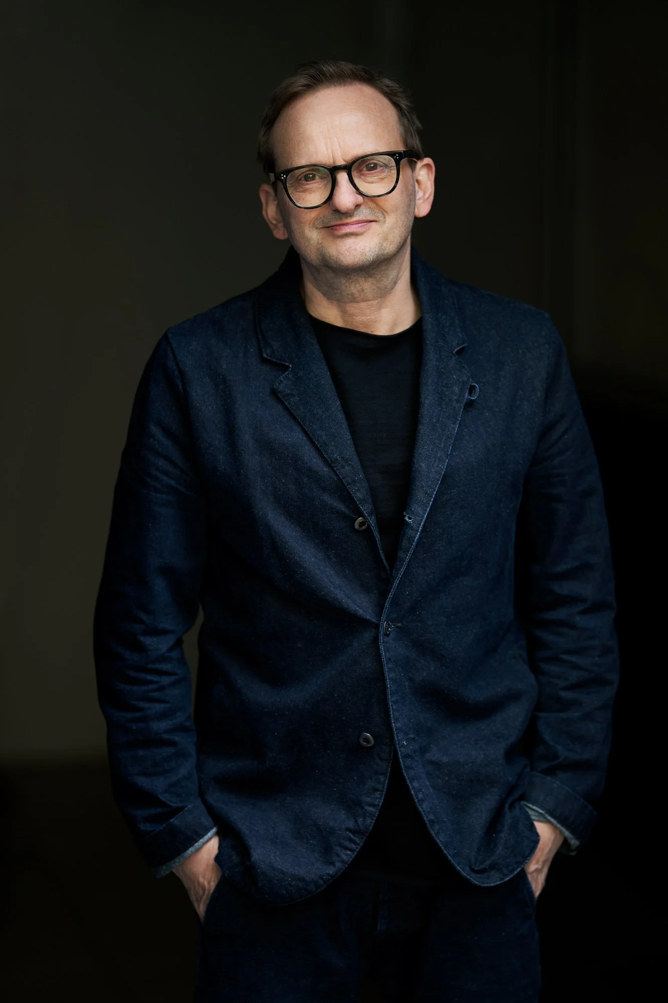 Porträtaufnahme des Schauspielers Milan Peschel mit Brille im dunkelblauen Jeansblazer und schwarzem Shirt vor dunklem Hintergrund, leichtes Lächeln, die Hände in den Taschen, fotografiert von Koszki Photography Berlin.