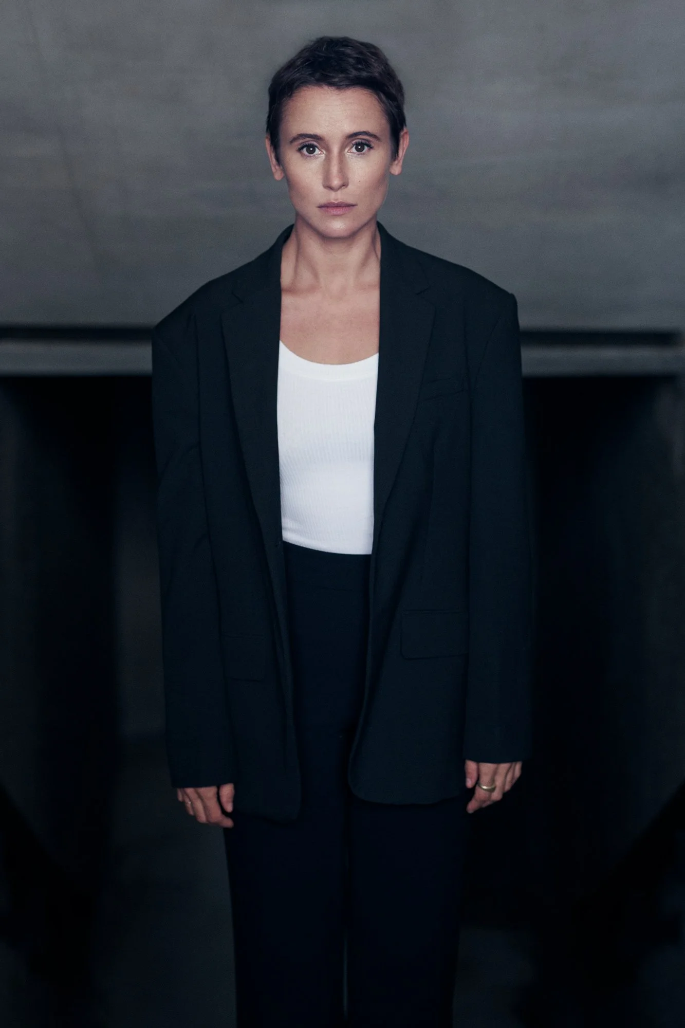 Porträtaufnahme der Schauspielerin Peri Baumeister im schwarzen Oversize-Blazer und weißem Top vor dunkler Betonwand, frontaler Blick in die Kamera, fotografiert von Koszki Photography Berlin.