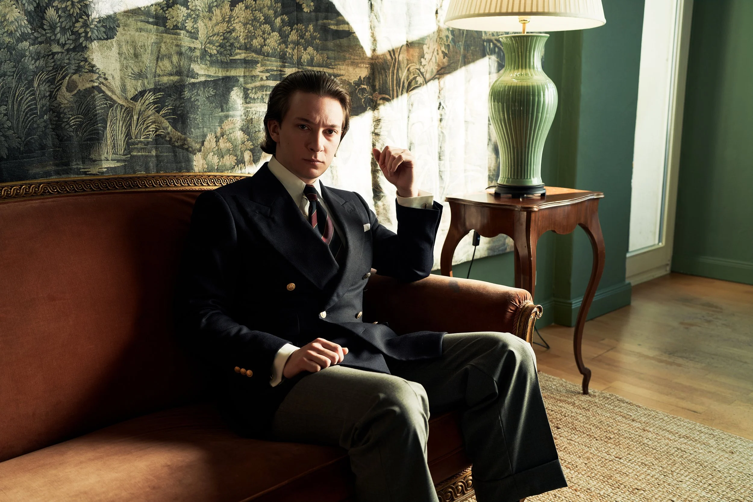 Ein elegant gekleideter Mann sitzt in einem klassisch eingerichteten Raum auf einem braunen Sofa. Er trägt ein dunkelblaues, doppelt geknöpftes Maßanfertigung-Sakko im Stil von Maximilian Mogg, kombiniert mit Krawatte und grauer Hose. Im Hintergrund 