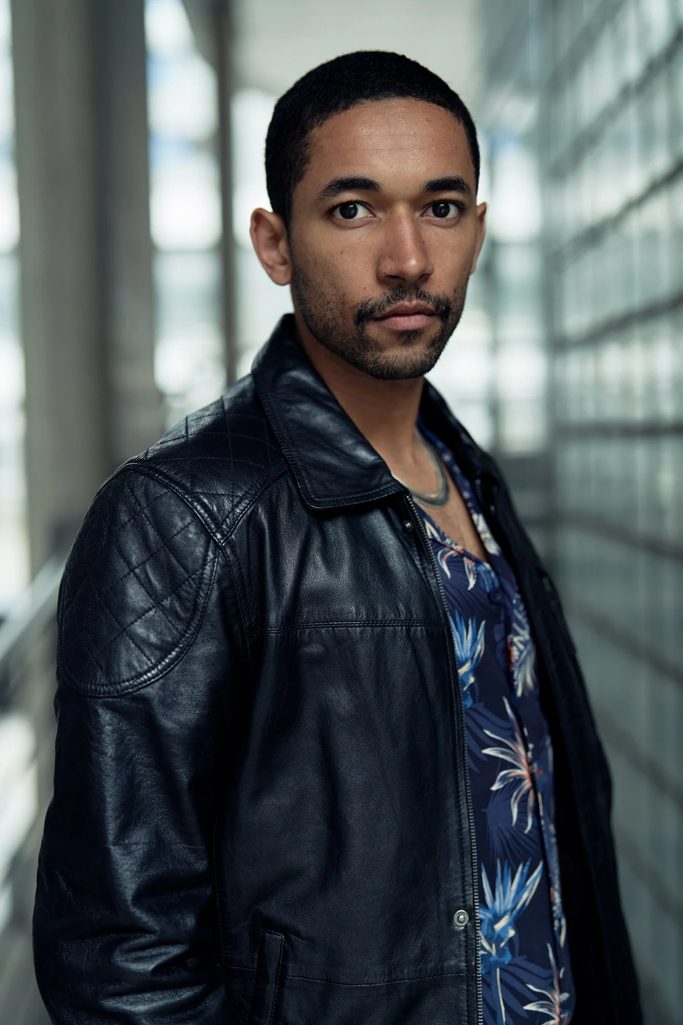 Alt-Text: Porträtaufnahme des Schauspielers Benito Bausel in schwarzer Lederjacke vor Glasfassade, direkter Blick in die Kamera, fotografiert von Koszki Photography Berlin.