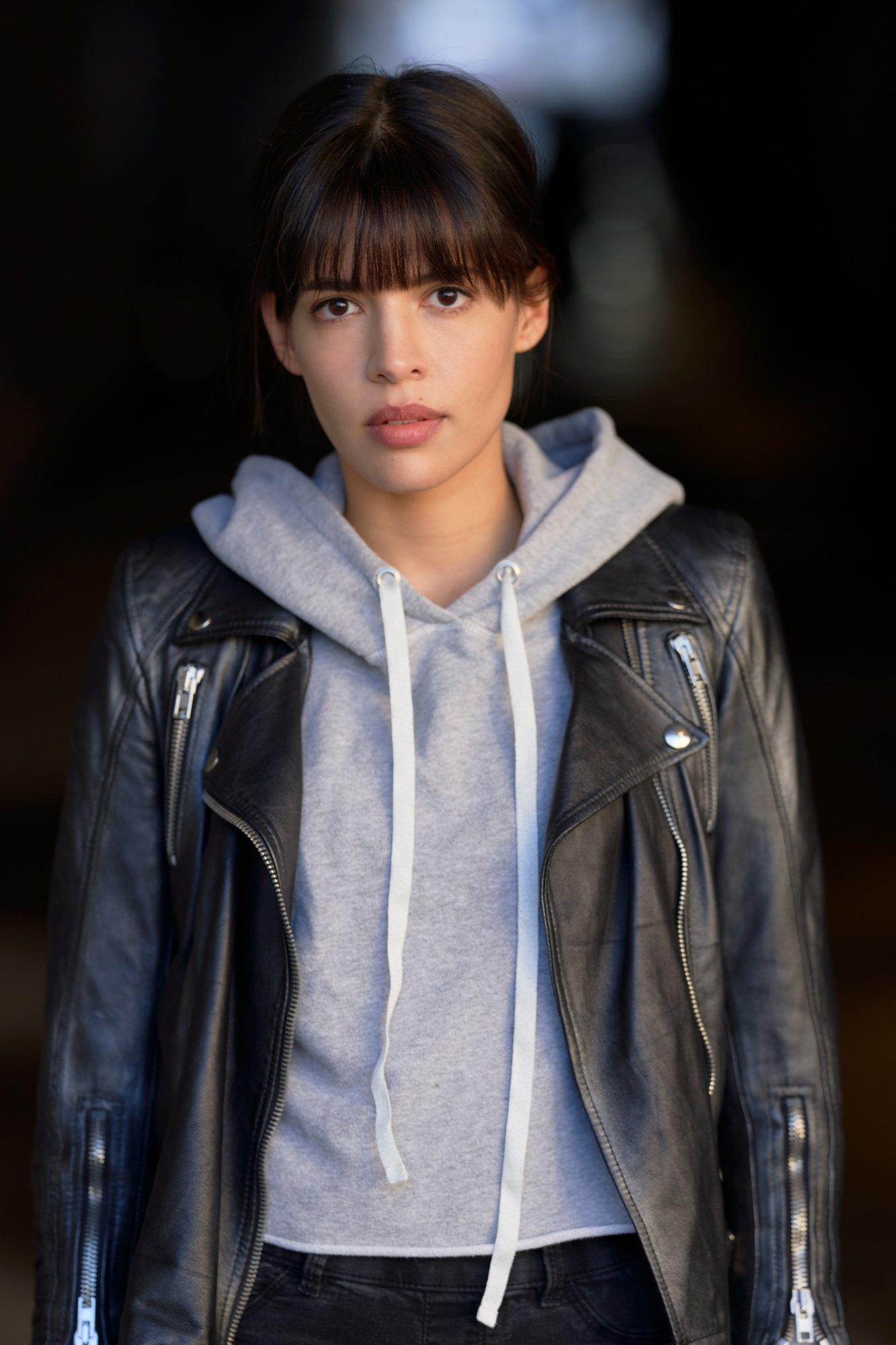 Porträt der Schauspielerin Jasmin Lord in schwarzer Lederjacke über grauem Kapuzenpullover mit direktem Blick in die Kamera vor dunklem Hintergrund, fotografiert von Koszki Photography Berlin.