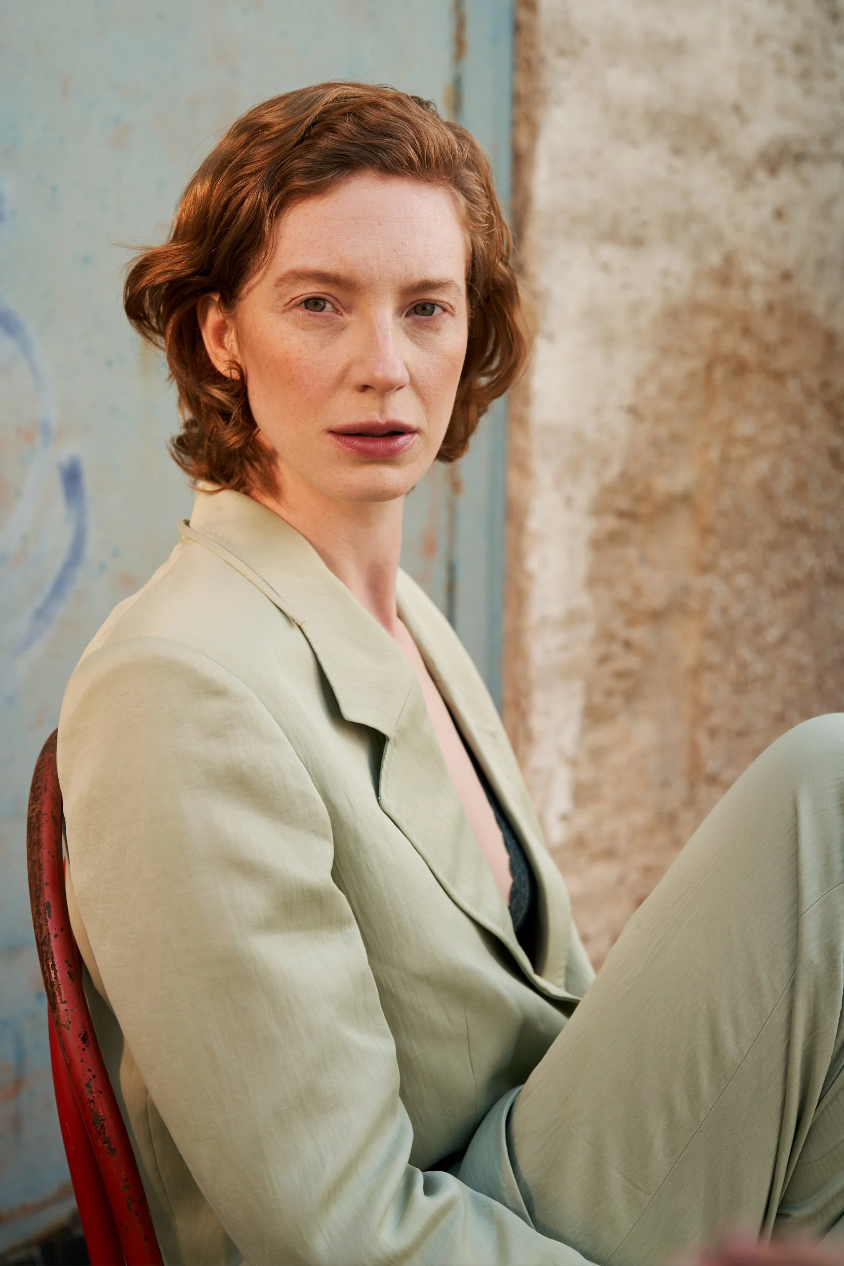 Porträt der Schauspielerin Luise Wolfram im hellgrünen Anzug auf einem roten Stuhl vor einer Putzwand, fotografiert von Koszki Photography Berlin.
