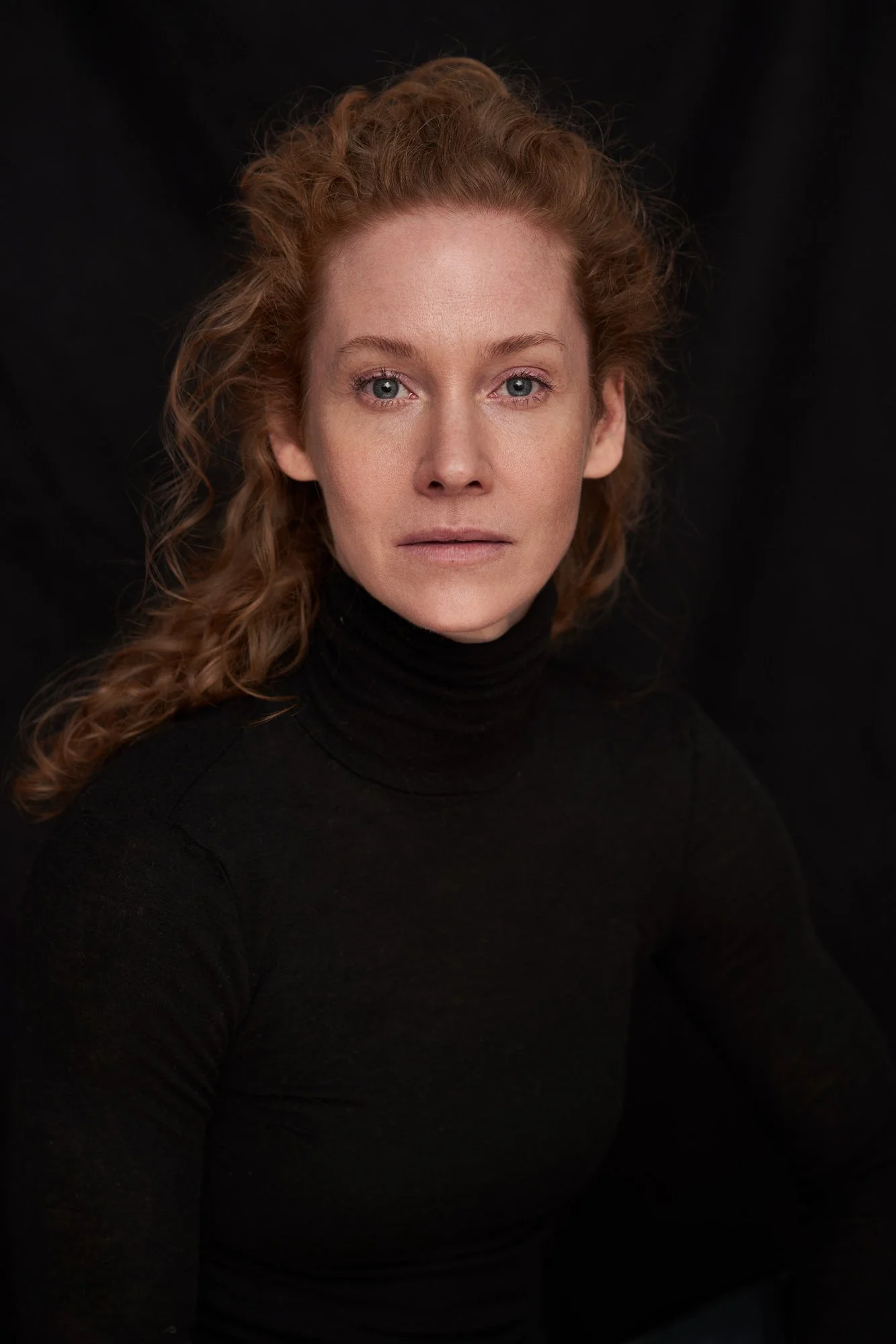 Headshot der Schauspielerin Sina-Valeska Jung mit lockigem rotem Haar im schwarzen Rollkragenpullover vor schwarzem Hintergrund, frontaler Blick in die Kamera, fotografiert von Koszki Photography Berlin.