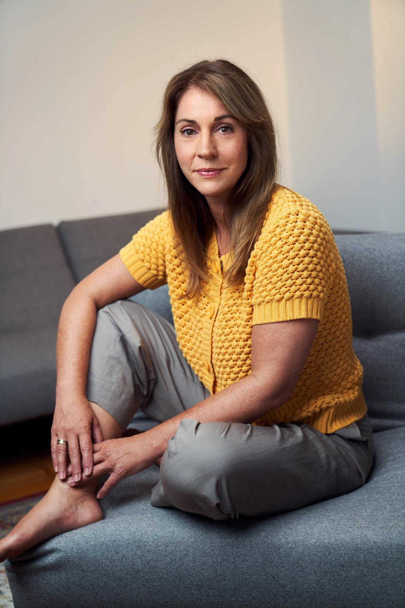 Porträtaufnahme der Schauspielerin Ulrike C. Tscharre in gelber Strickjacke, entspannt auf einem grauen Sofa sitzend, leichtes Lächeln, fotografiert von Koszki Photography Berlin.