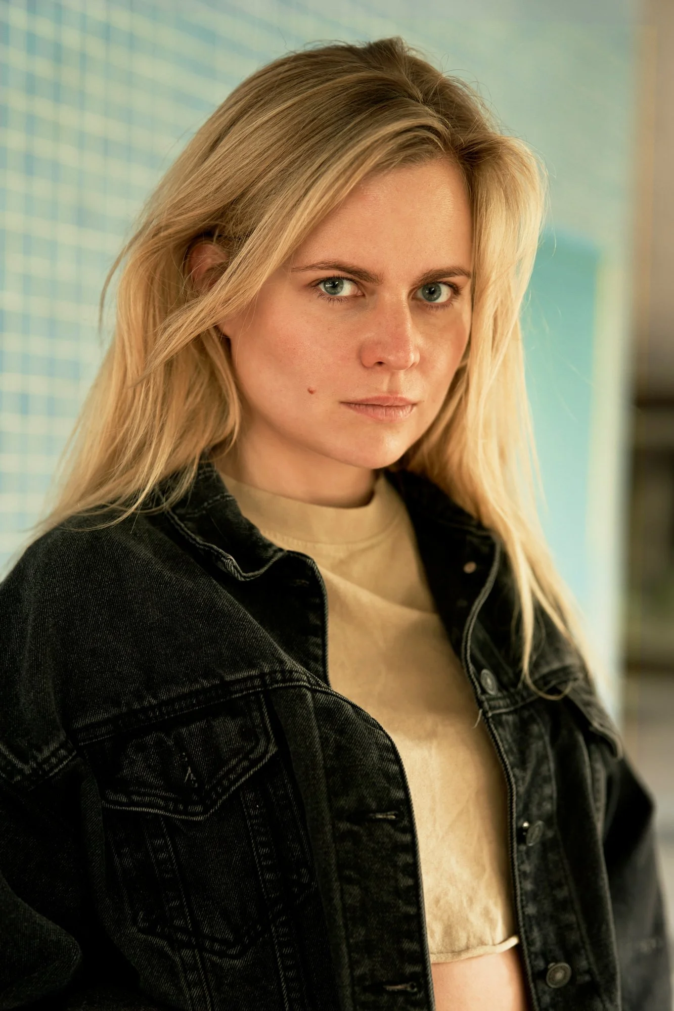 Headshot der Schauspielerin und Sängerin Kara Edenberg mit langen blonden Haaren in schwarzer Jeansjacke über beigem Top, mit intensivem Blick in die Kamera vor einer blauen Mosaikwand, fotografiert von Koszki Photography Berlin.