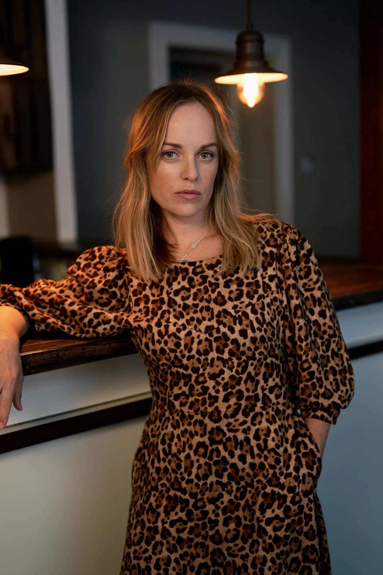 orträtaufnahme der Schauspielerin Friederike Kempter im Kleid mit Leopardenmuster, an einen Holztresen gelehnt, im Hintergrund eine warme Pendelleuchte, fotografiert von Koszki Photography Berlin.
