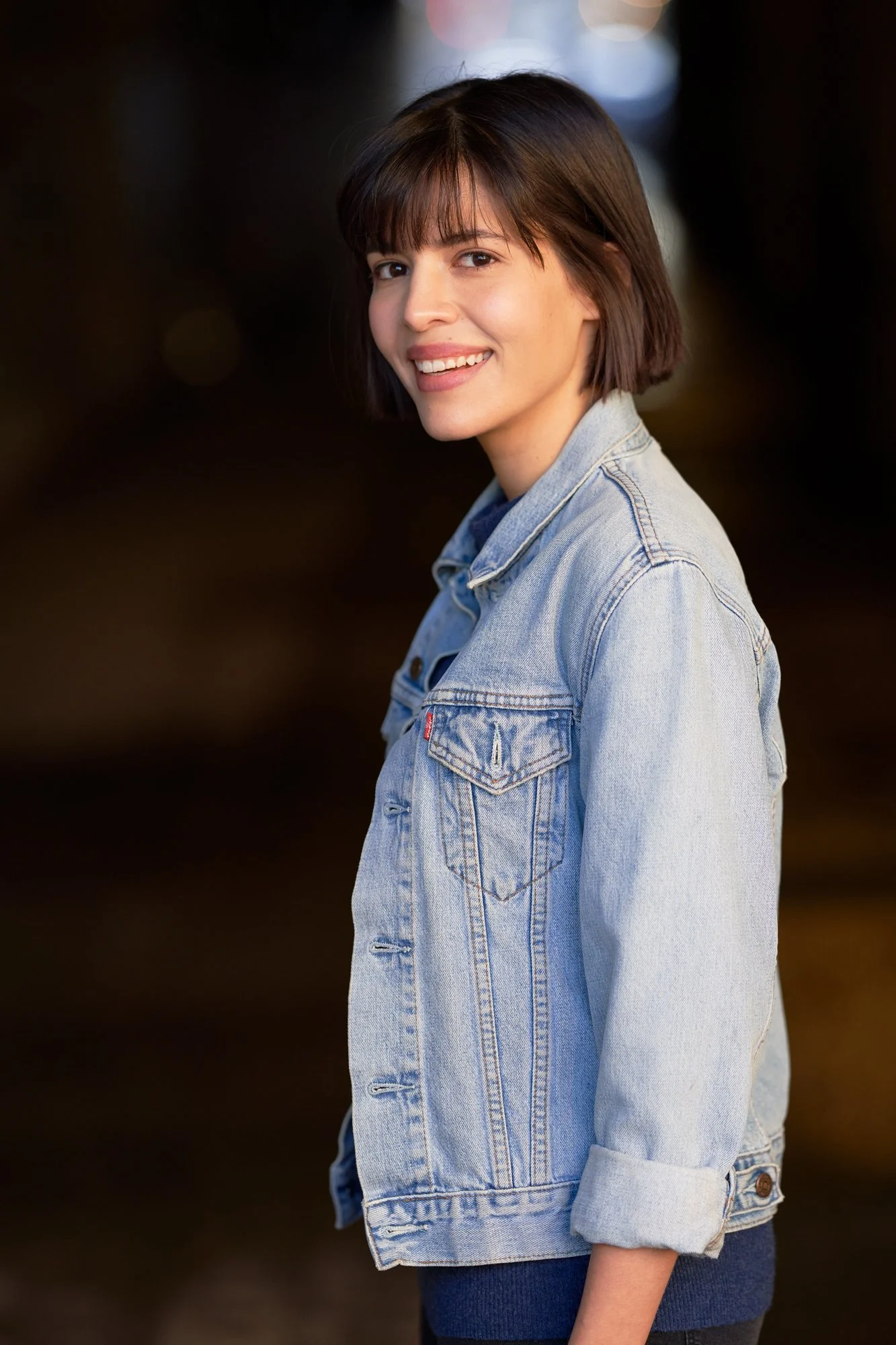 Porträt der lächelnden Schauspielerin Jasmin Lord in heller Jeansjacke, seitlich stehend mit Blick über die Schulter in die Kamera vor dunklem Hintergrund, fotografiert von Koszki Photography Berlin.