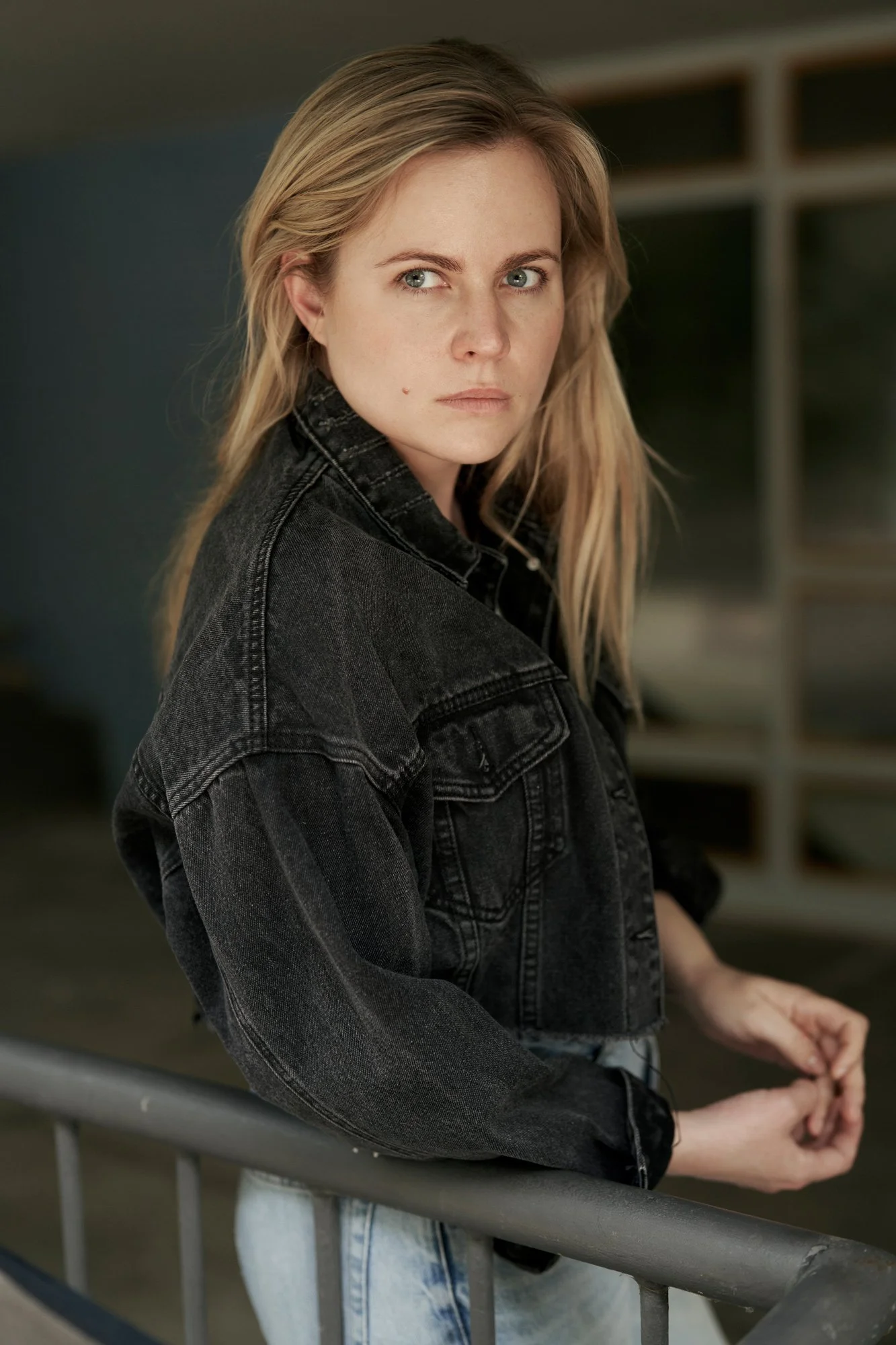 Porträt der Schauspielerin und Sängerin Kara Edenberg mit langen blonden Haaren in schwarzer Jeansjacke, an ein Metallgeländer gelehnt, mit ernstem Blick über die Schulter in die Kamera, fotografiert von Koszki Photography Berlin.