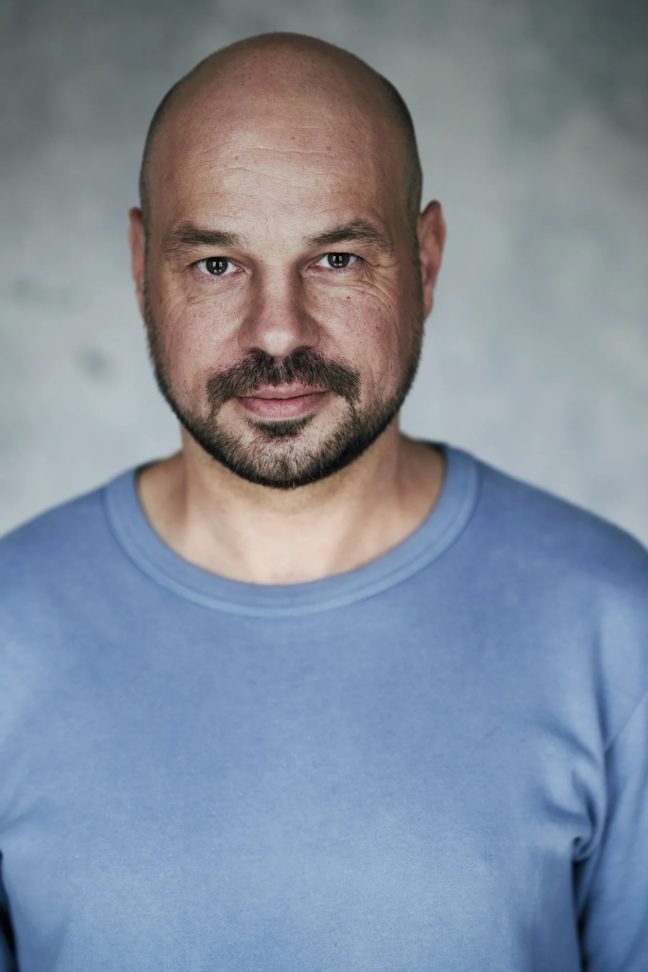 Farbporträt des DJs und Produzenten D-Nox im hellblauen Pullover mit ruhigem, direktem Blick vor grauem Betonhintergrund, fotografiert von Koszki Photography Berlin.