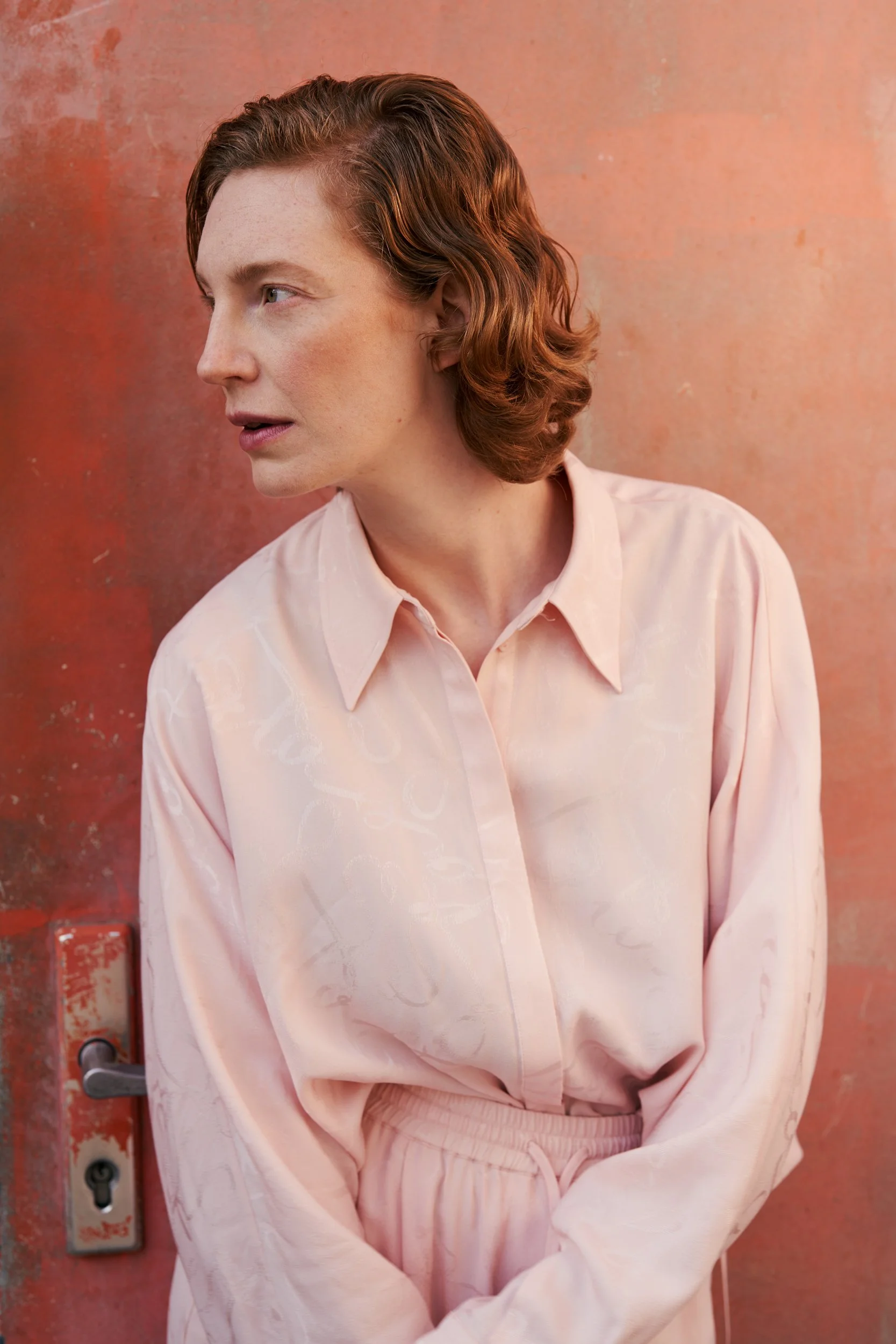 Porträt der Schauspielerin Luise Wolfram in rosa Bluse vor einer rostigen blaugrauen Metallwand mit direktem Blick, fotografiert von Koszki Photography Berlin.