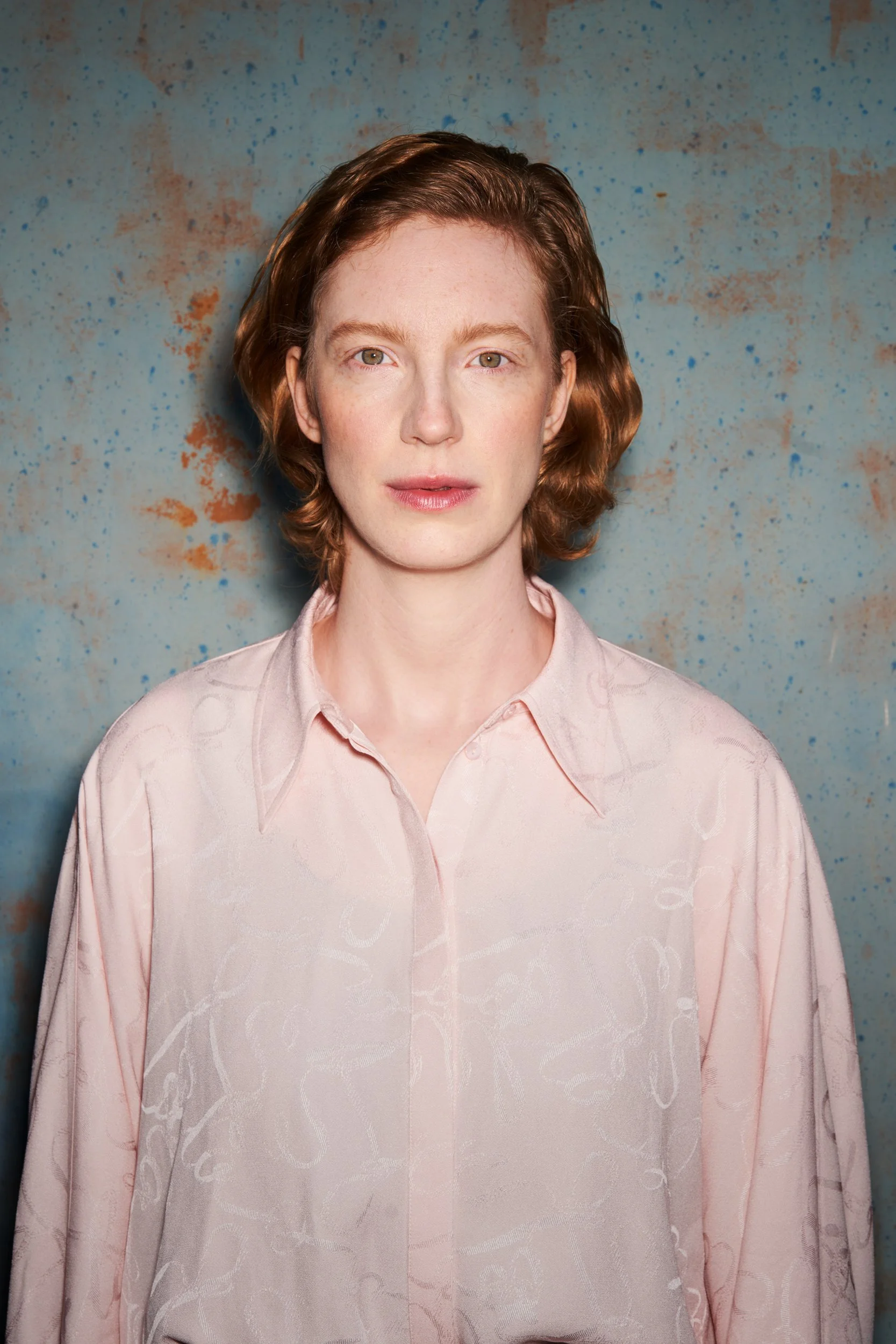 Porträt der Schauspielerin Luise Wolfram in rosa Bluse vor einer rostigen blaugrauen Metallwand mit direktem Blick, fotografiert von Koszki Photography Berlin.