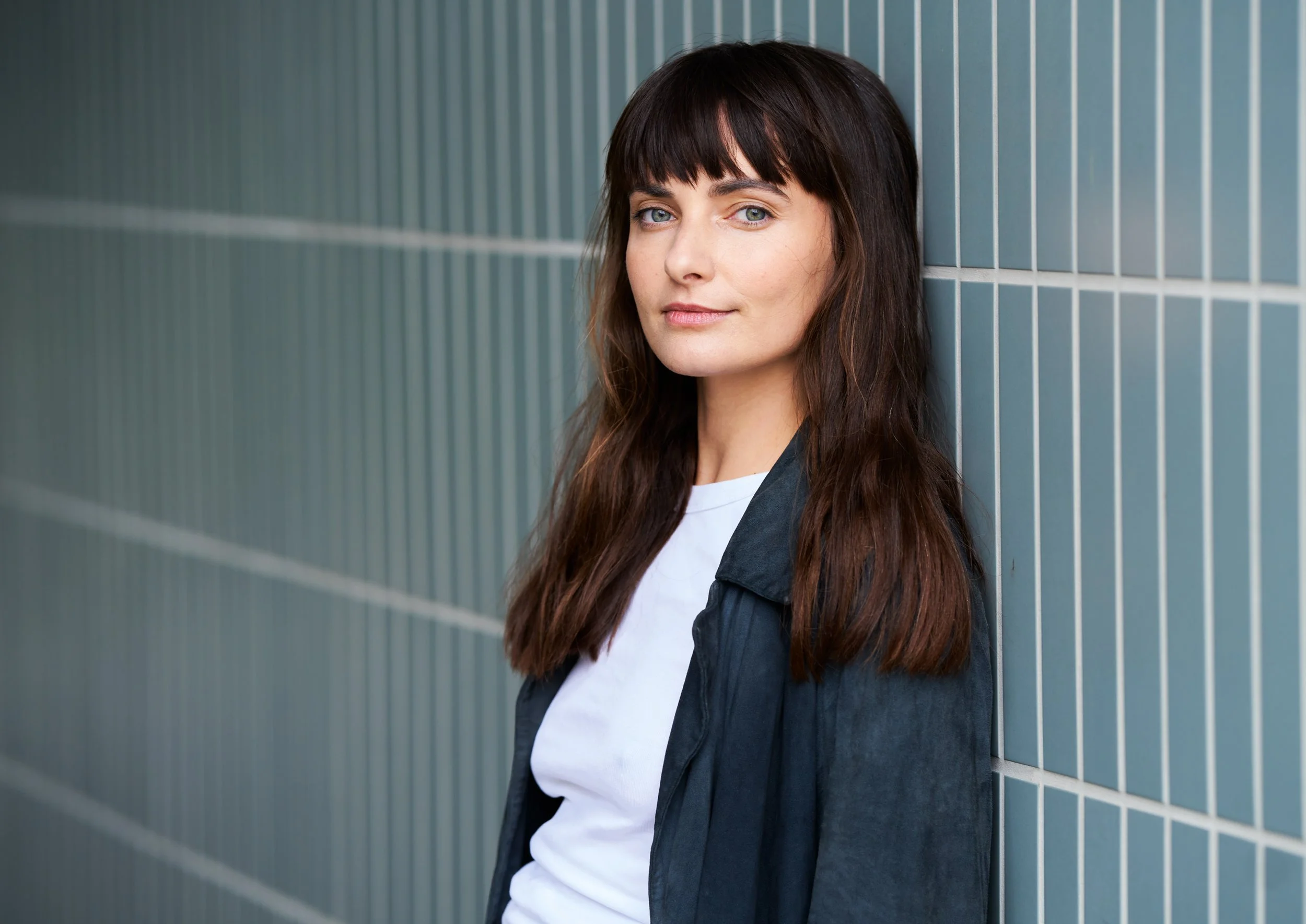 Porträtaufnahme der Schauspielerin Paula Schramm mit langem dunklem Haar und Pony in dunkler Wildlederjacke und weißem Shirt, an eine blaugrüne Fliesenwand gelehnt, leichtes Lächeln, fotografiert von Koszki Photography Berlin.